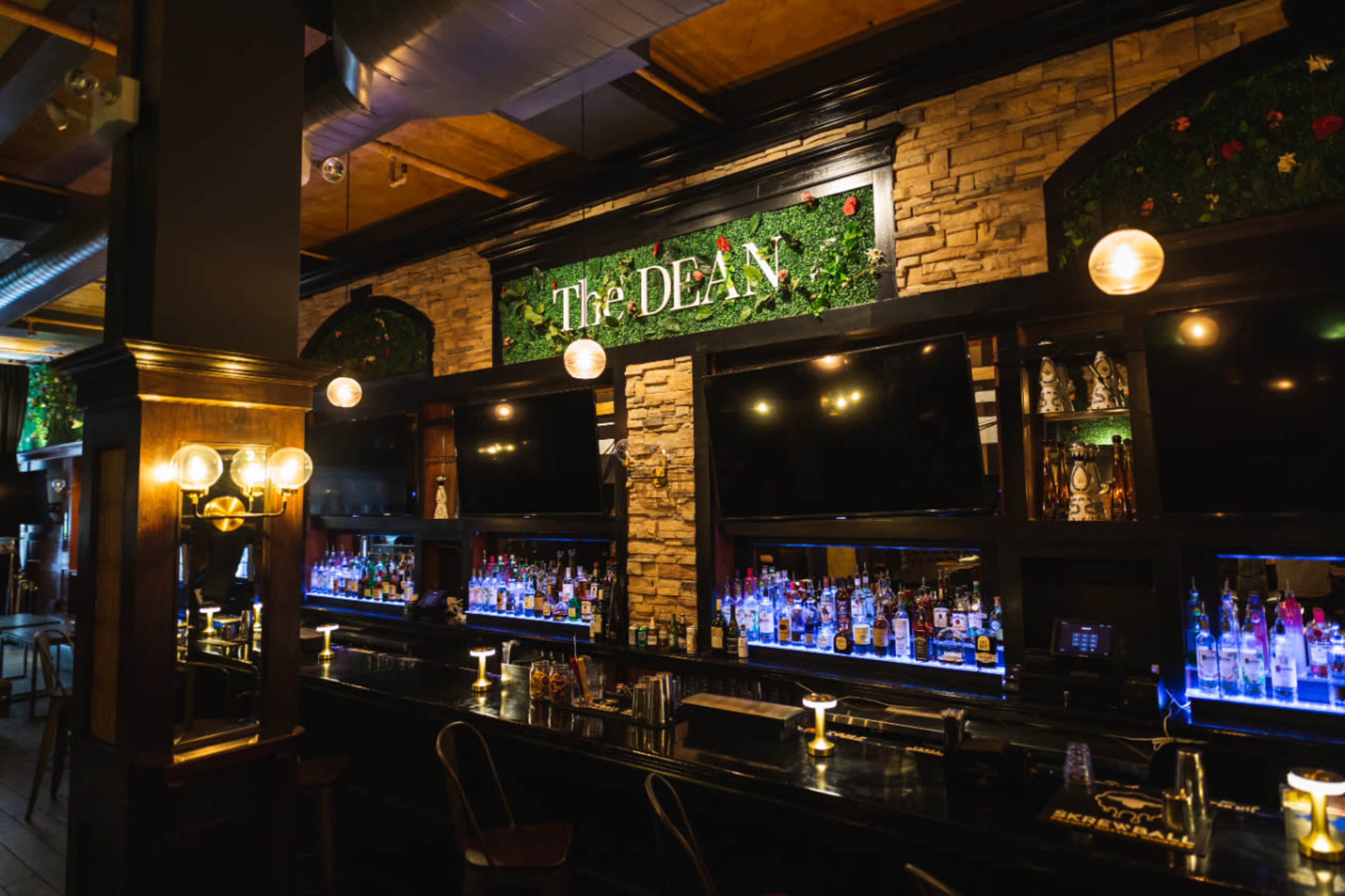 The image shows a well-lit bar with a wooden counter, shelves of various drinks, and decorative greenery above the signage that reads "The DEAN."