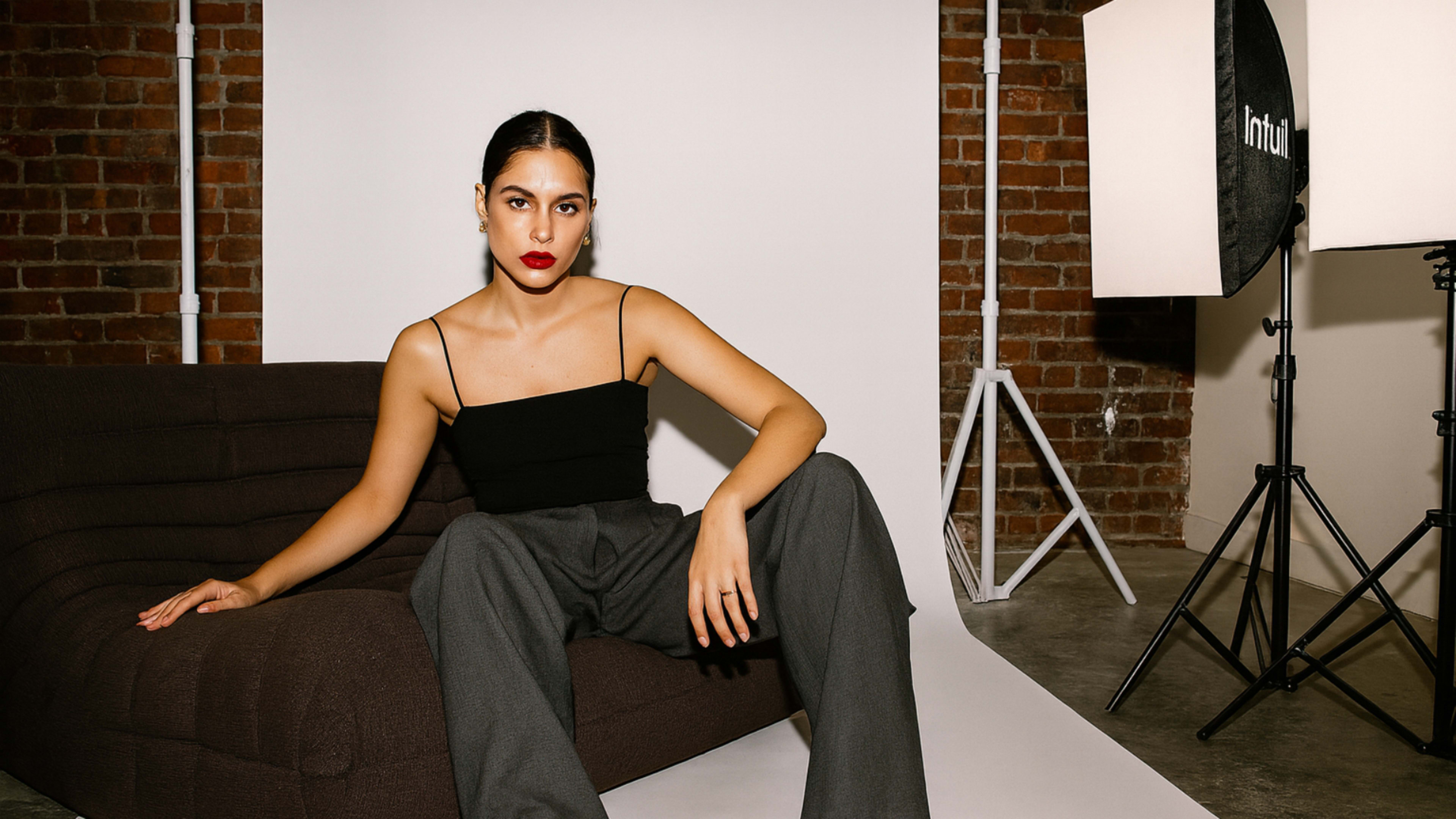 A person sits on a brown couch in front of a backdrop, wearing a black top and gray pants, with studio lighting and brick walls in the background.