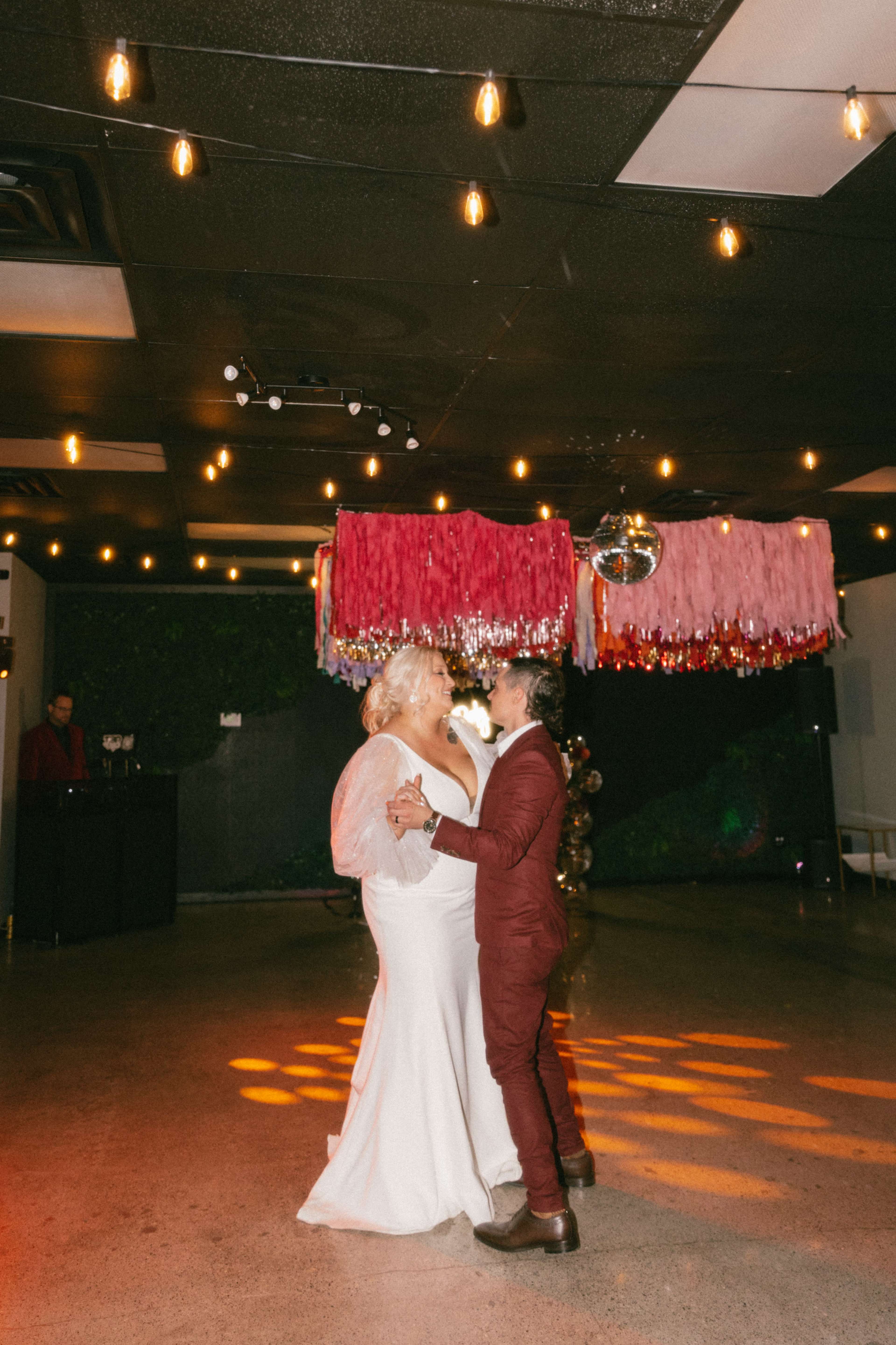 A couple dances together in a dimly lit venue with colorful decor overhead and string lights illuminating the space.