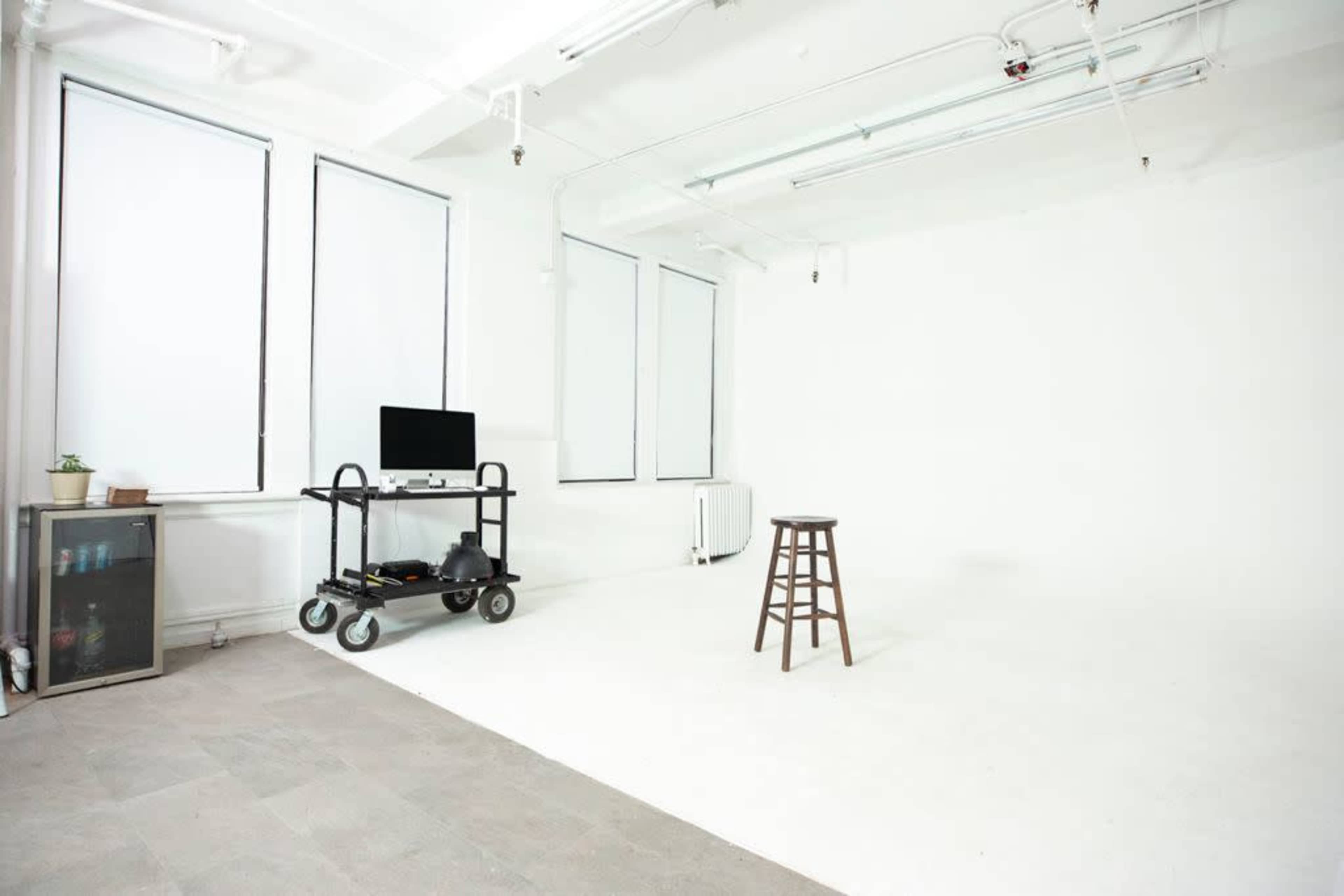 The image shows an empty studio space with white walls, a wooden stool in the center, a cart with a computer, and large windows letting in natural light.