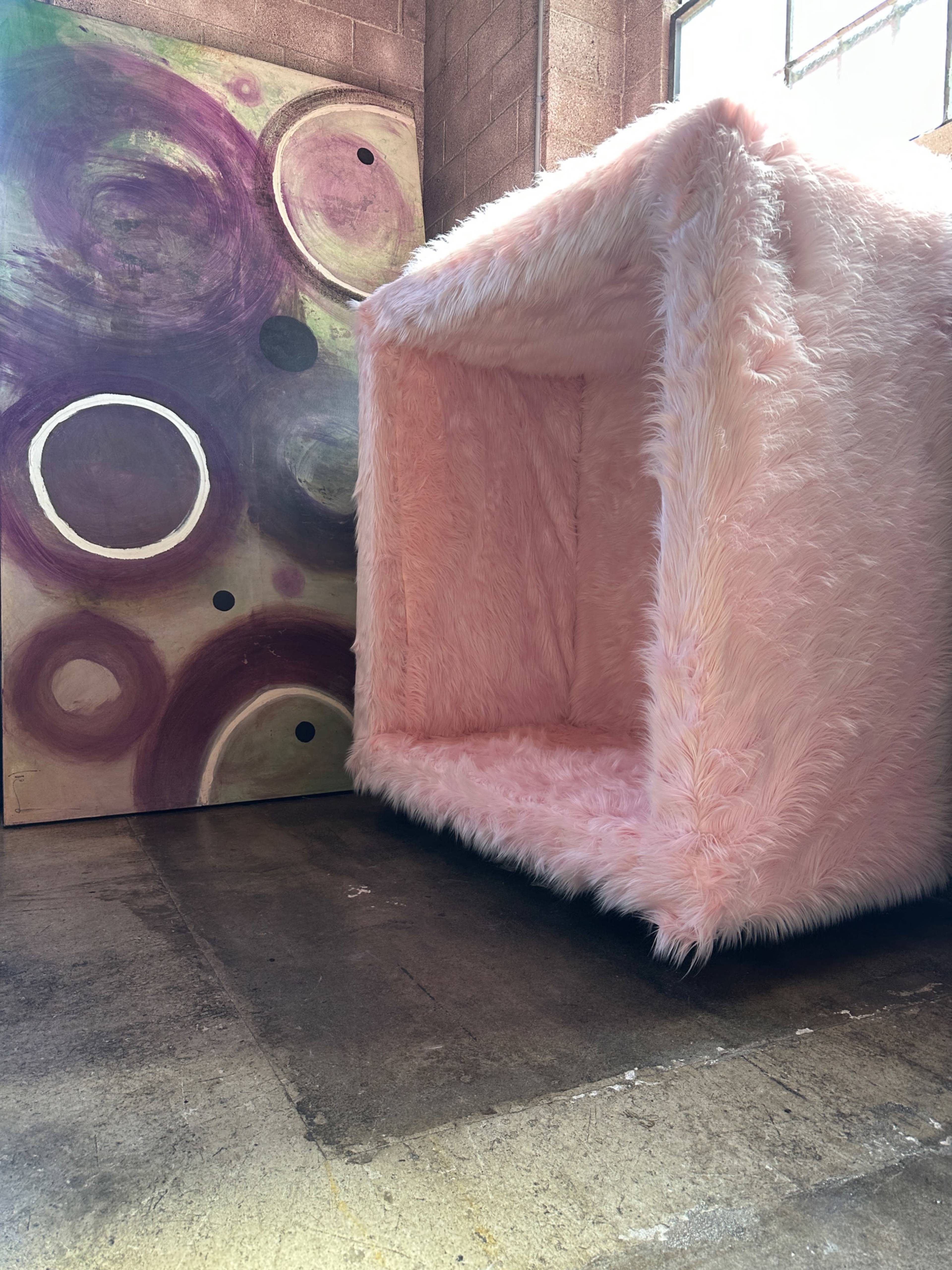 A large, pink furry cube structure is positioned next to a colorful abstract mural with circular patterns on a concrete floor.