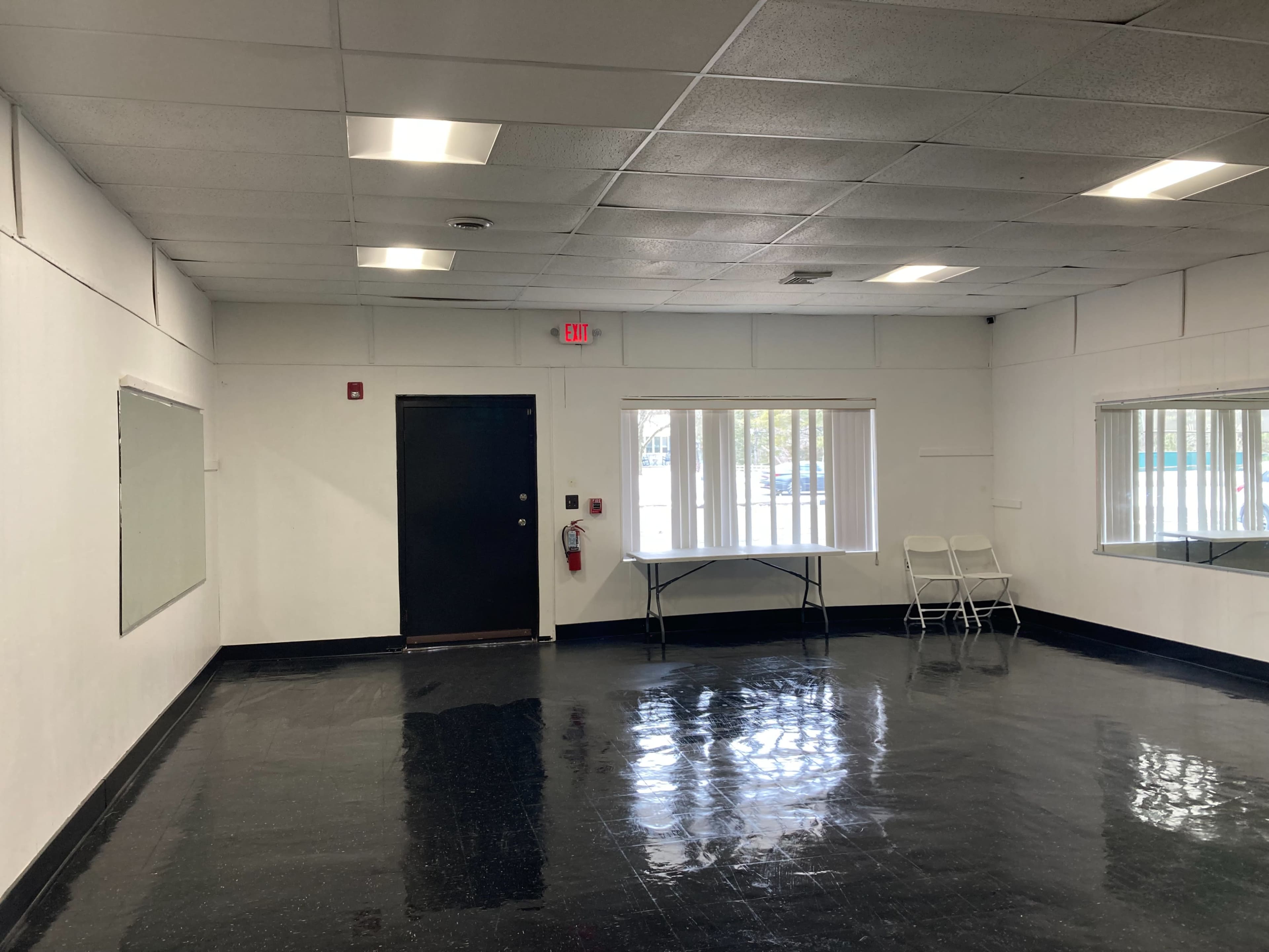 A sparsely furnished room with a black floor, a table with chairs, and two windows allowing natural light.