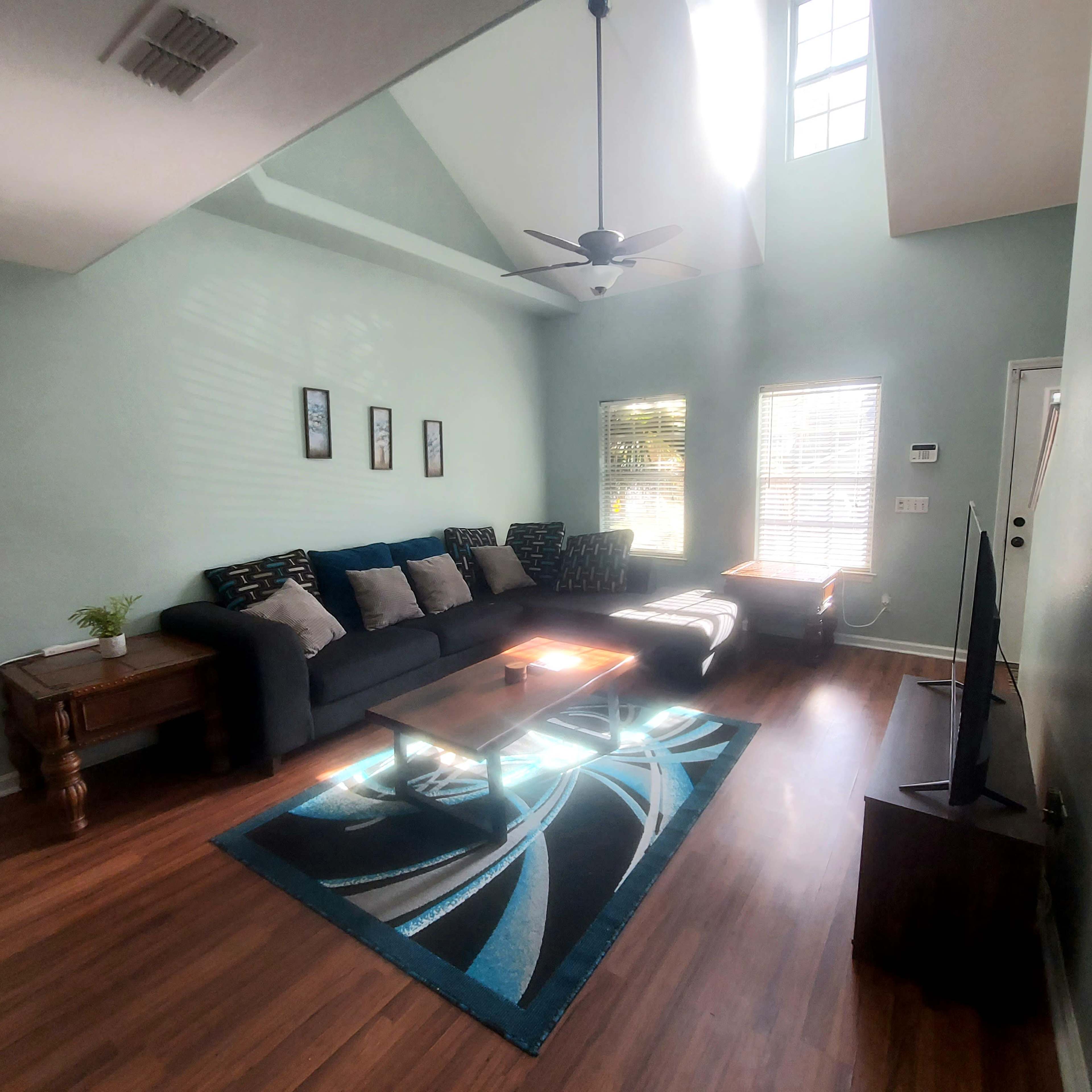 A living room with a sectional couch, a coffee table on a patterned rug, and large windows allowing natural light.