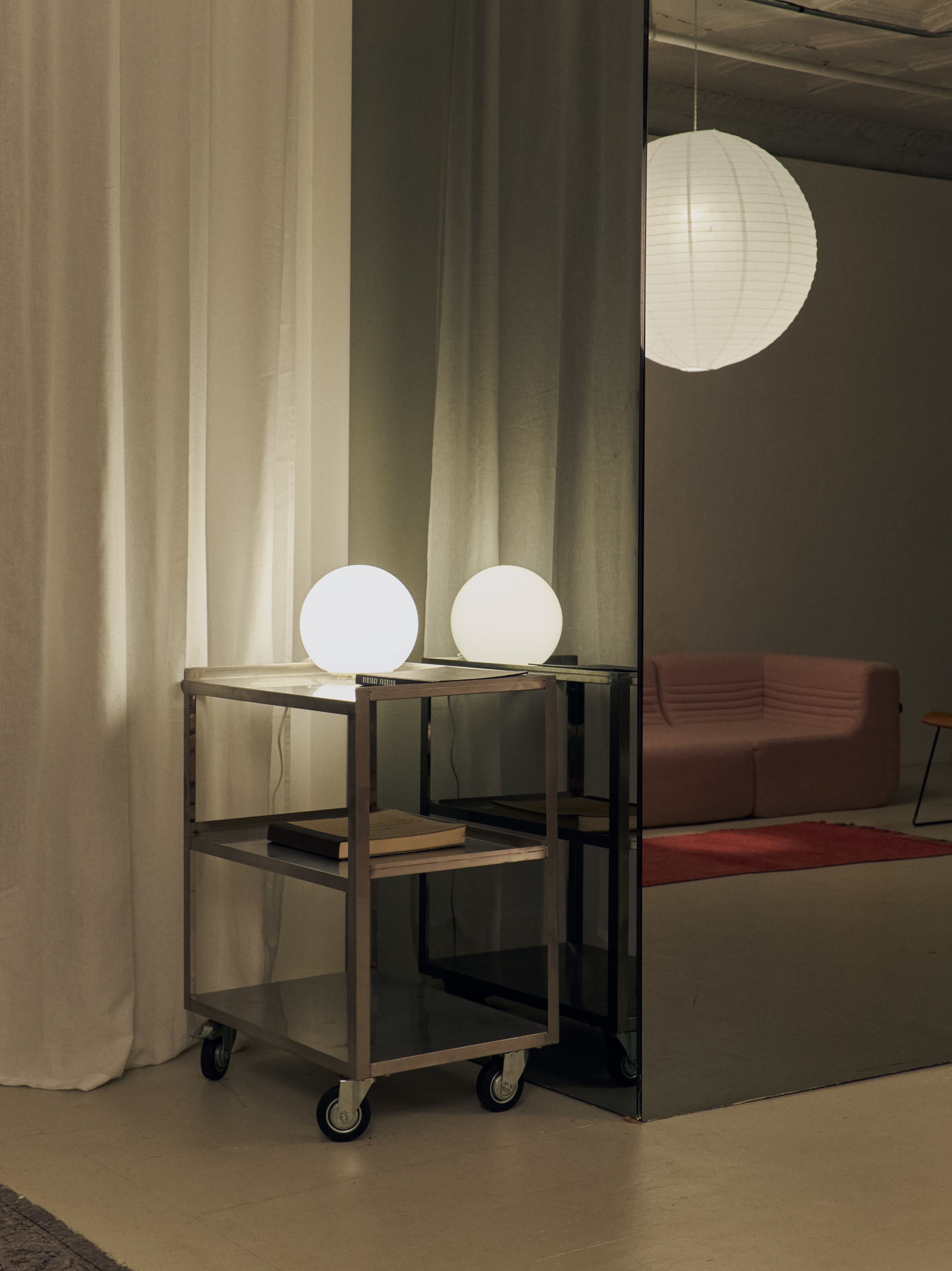 A metal cart with two spherical lamps sits beside a mirror, reflected in a modern interior space with soft lighting.