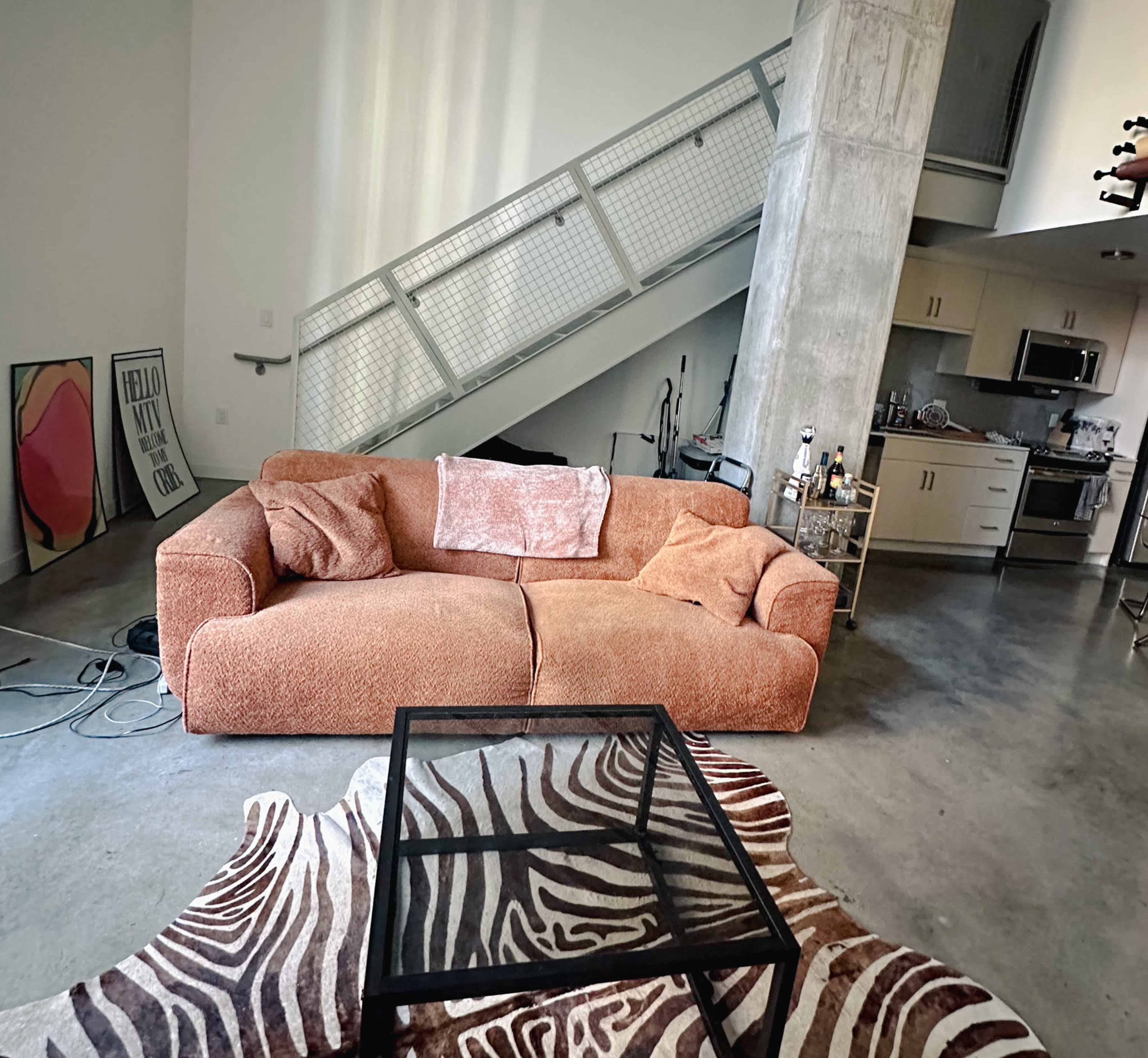 The image shows a spacious living room featuring a large orange sofa, a glass coffee table with a zebra print rug, and a staircase in the background leading to an upper level.