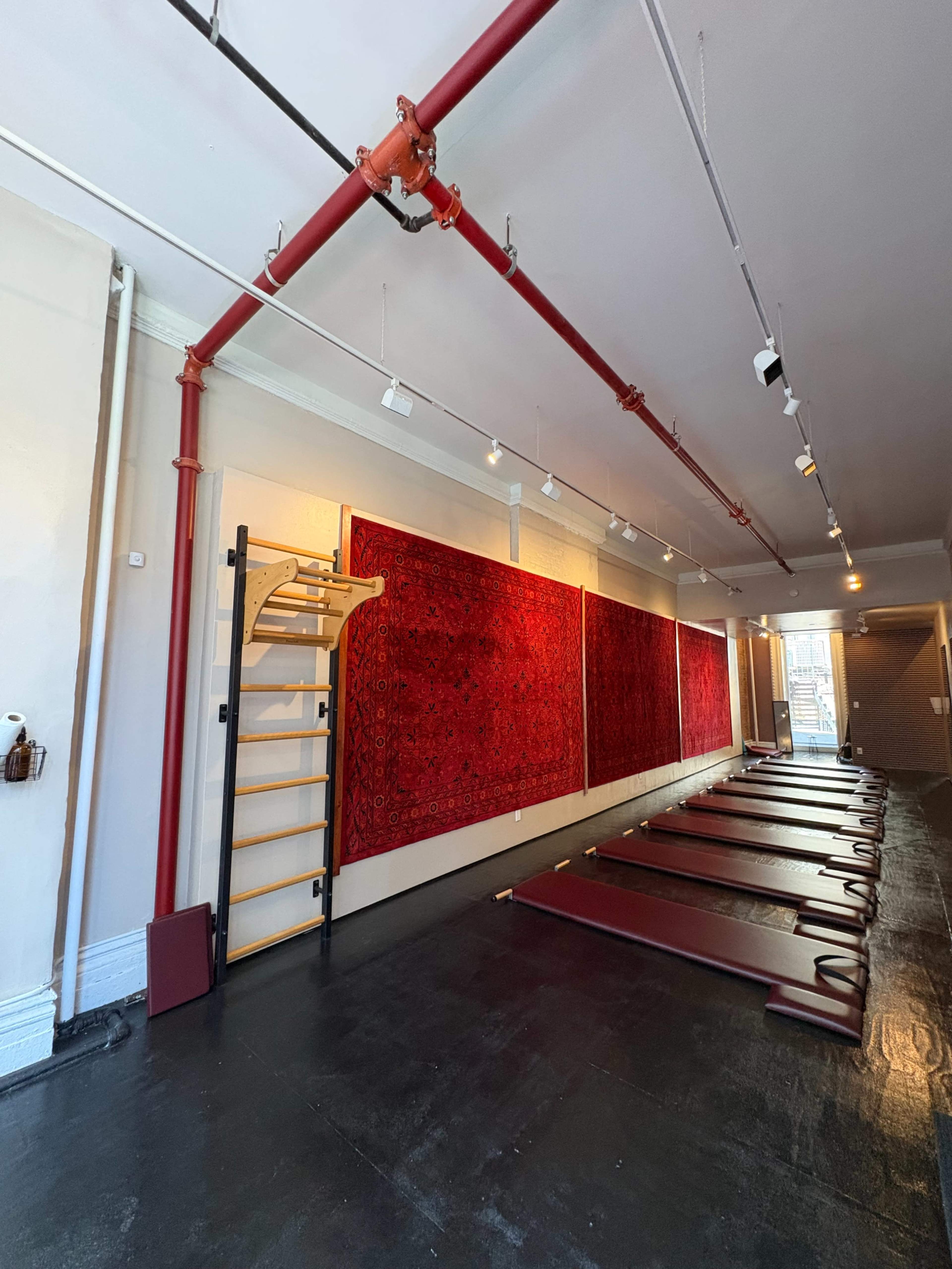 The image shows a spacious room with red exercise mats arranged on the floor and large red carpets hanging on the wall, along with exposed pipes and overhead lighting.