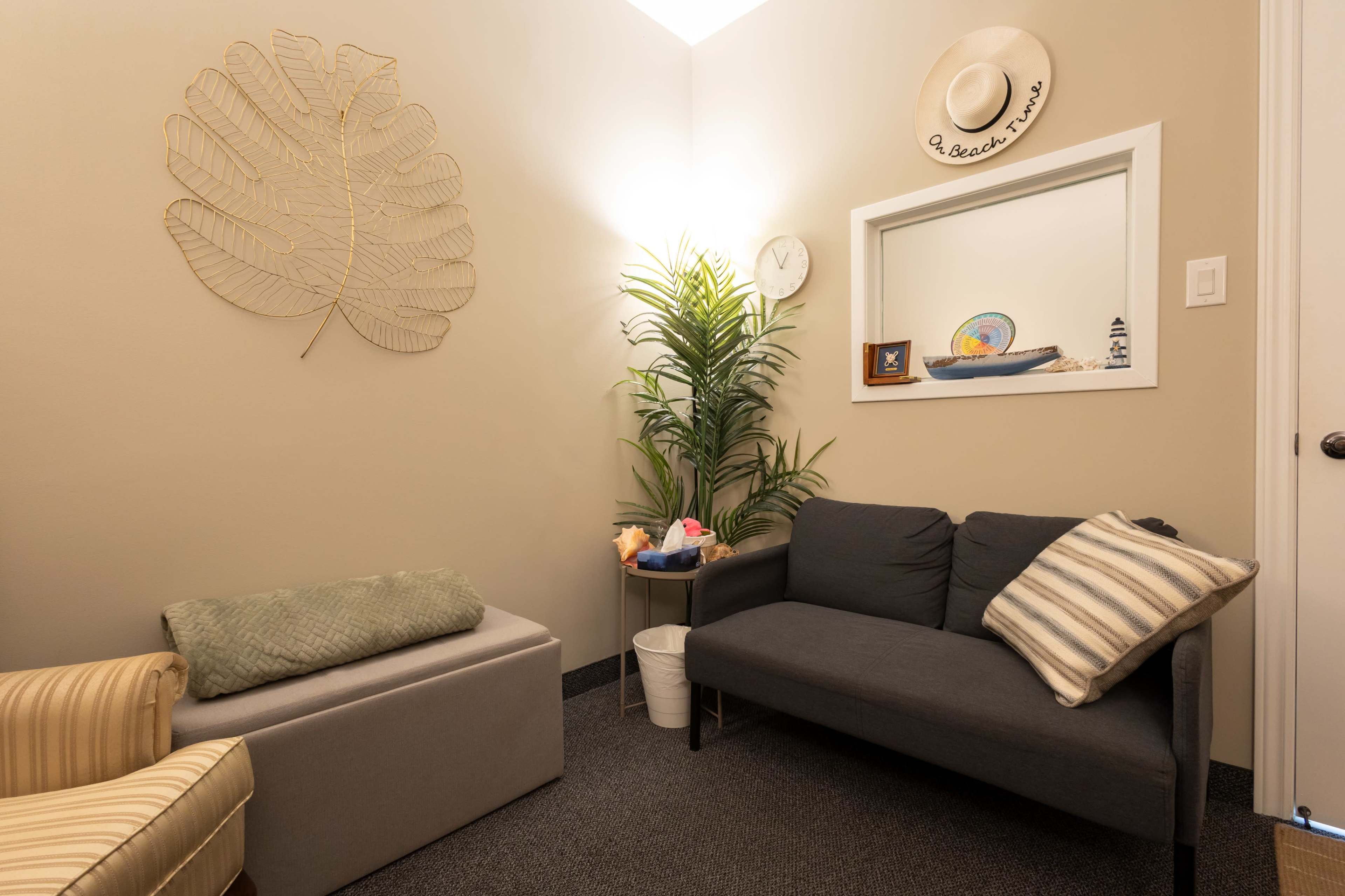The image shows a cozy room with a gray sofa, a light-colored chair, a potted plant, and decorative items on the walls and a shelf.