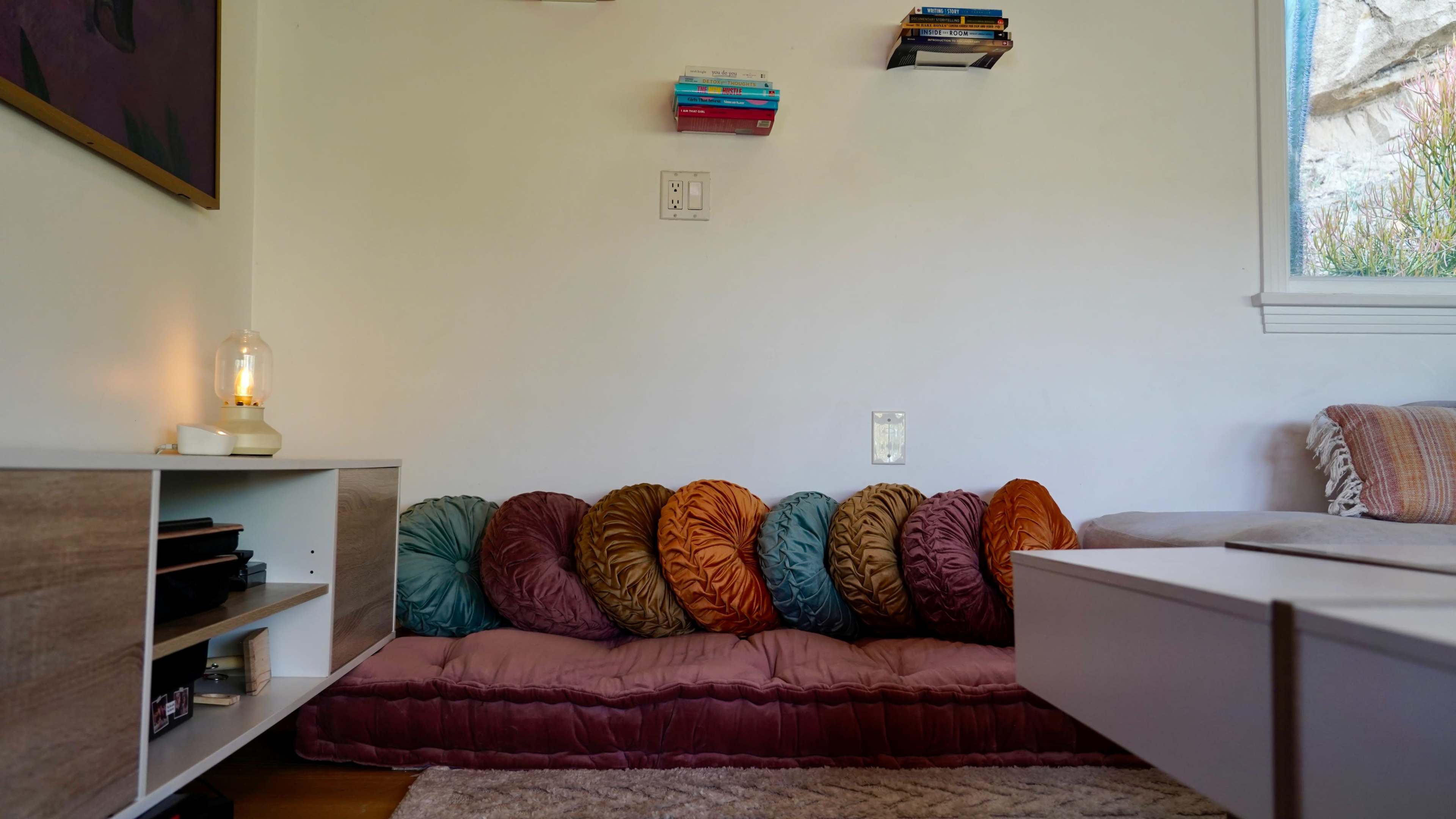 The image shows a cozy seating area with colorful round pillows arranged on a low couch, accompanied by a small bookshelf mounted on the wall.