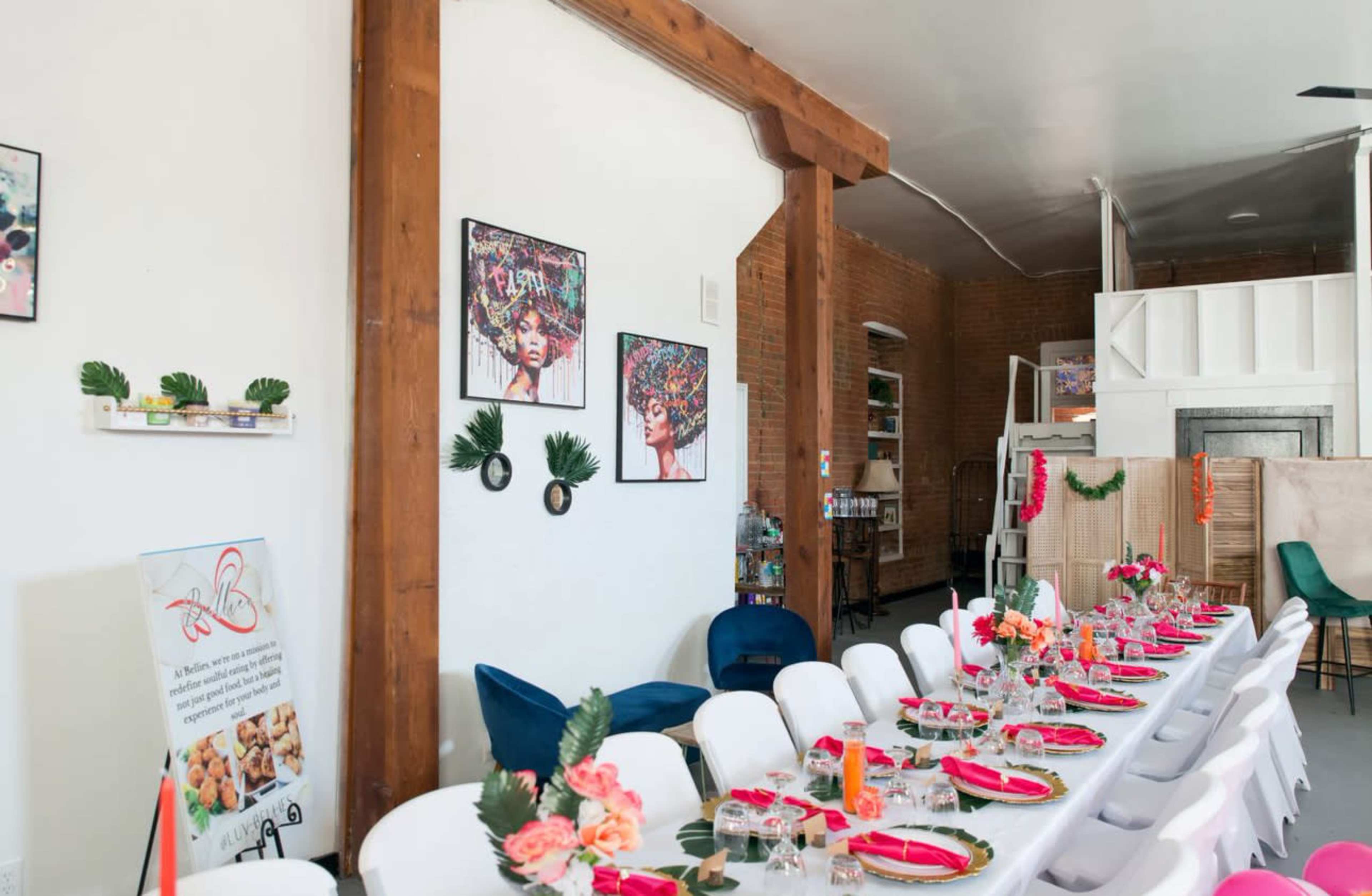 A long dining table is set with pink napkins and flowers, surrounded by chairs in a room featuring exposed wooden beams and colorful artwork on the walls.