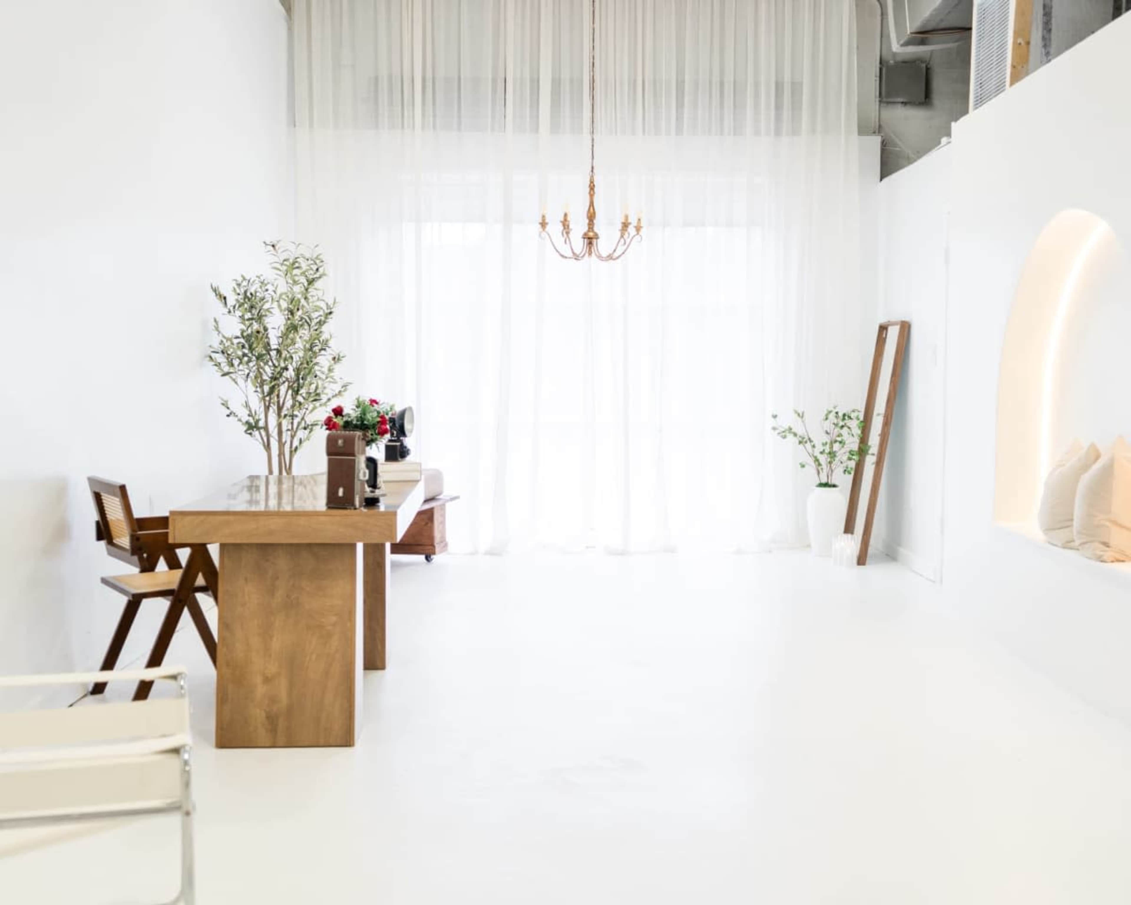 The image shows a minimalist white room featuring a wooden table, a chair, and large windows covered by sheer curtains.