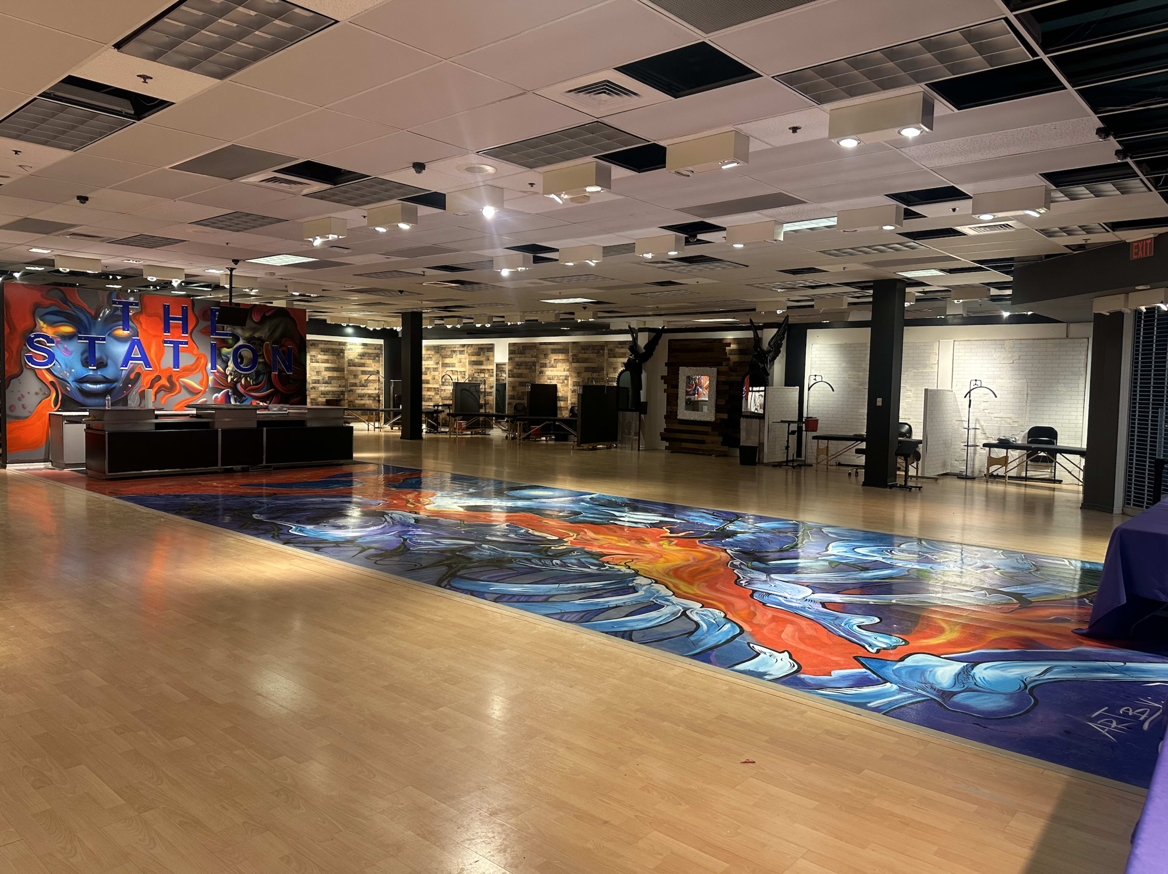 The image shows a spacious indoor venue featuring a colorful mural on the floor and an artistic backdrop with various seating areas.