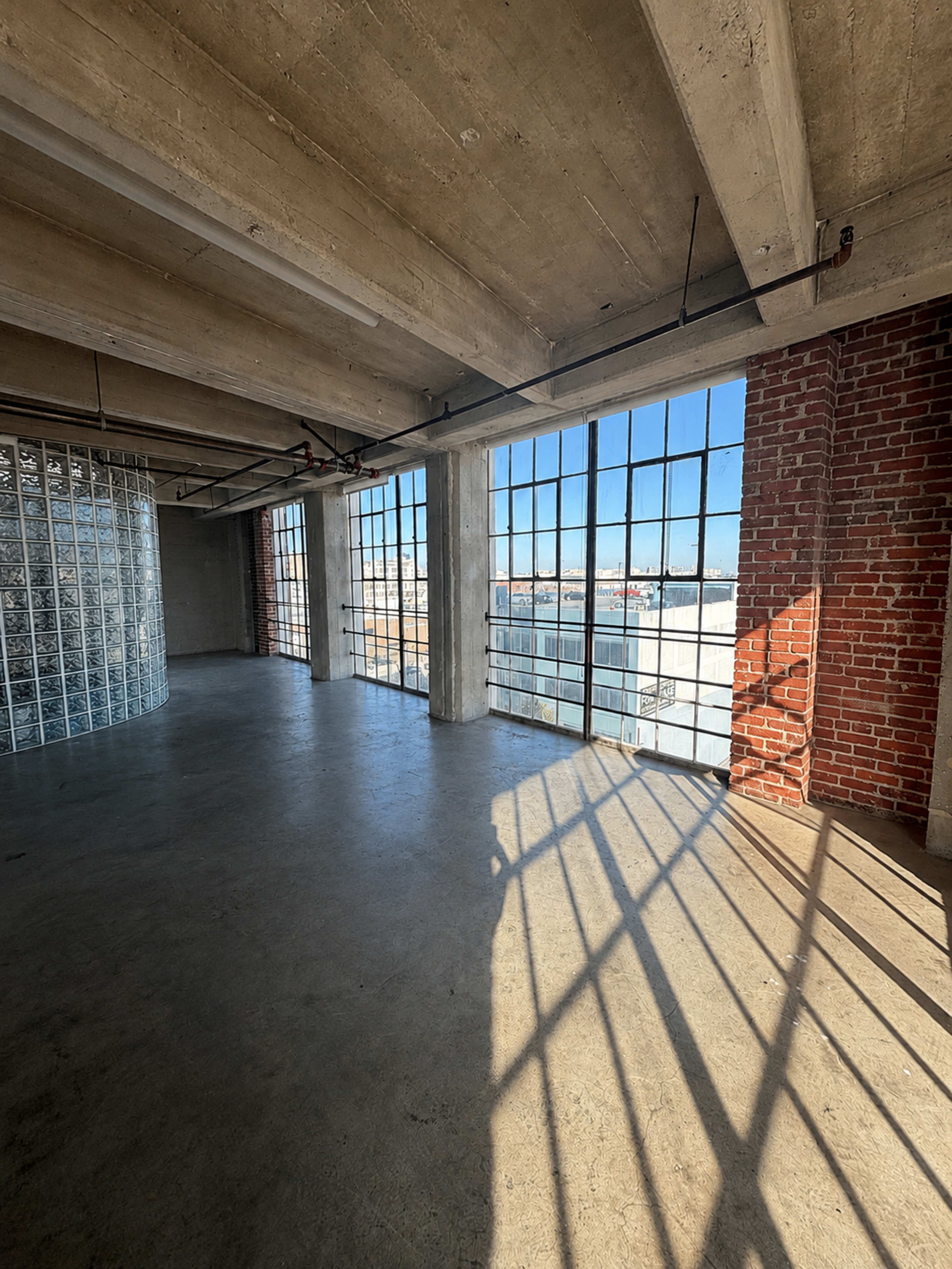 The image shows an empty industrial-style interior with large windows allowing natural light and casting shadows on the concrete floor.
