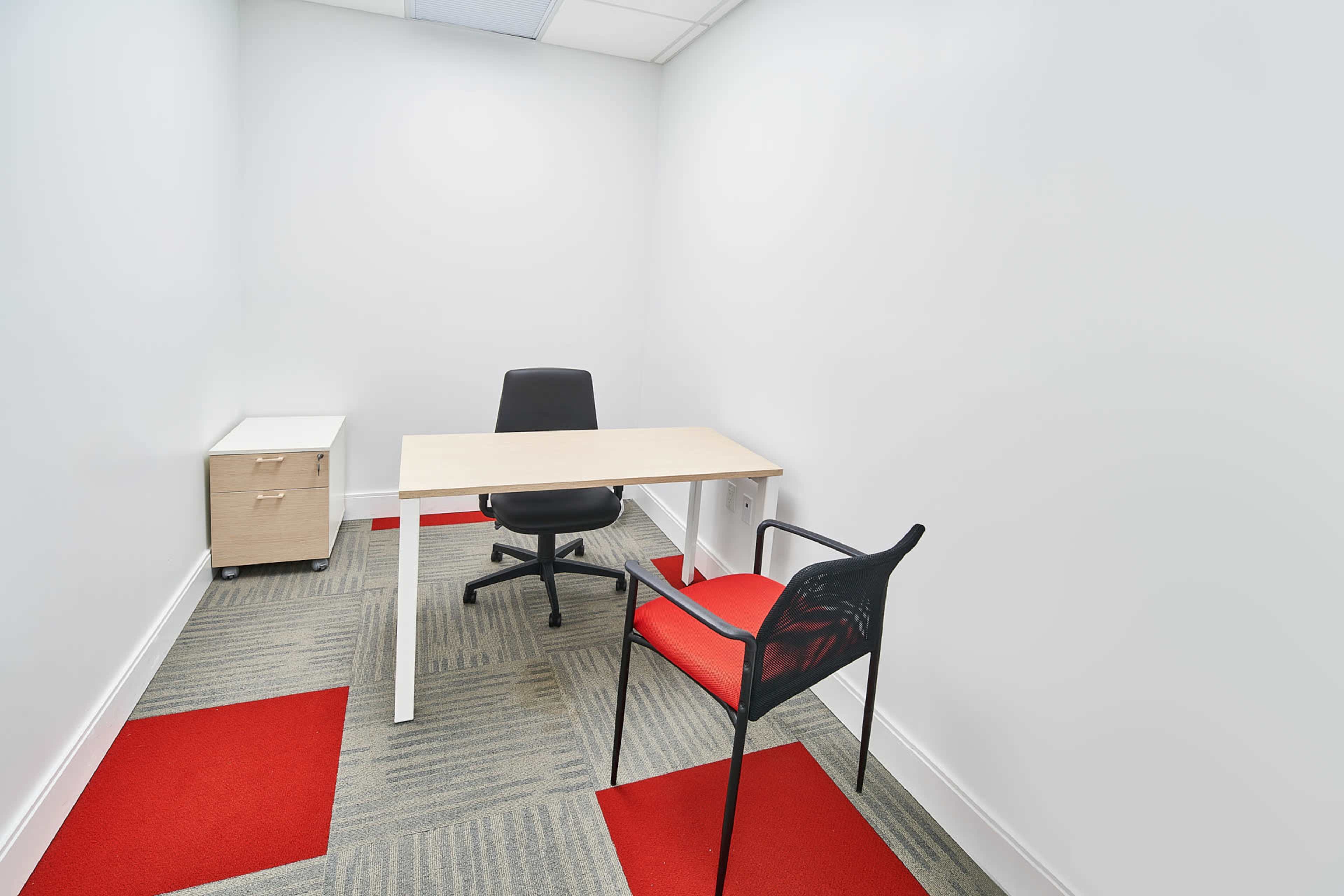 A small, empty room with a desk, a chair, and a storage unit, featuring a gray carpet with red square tiles.