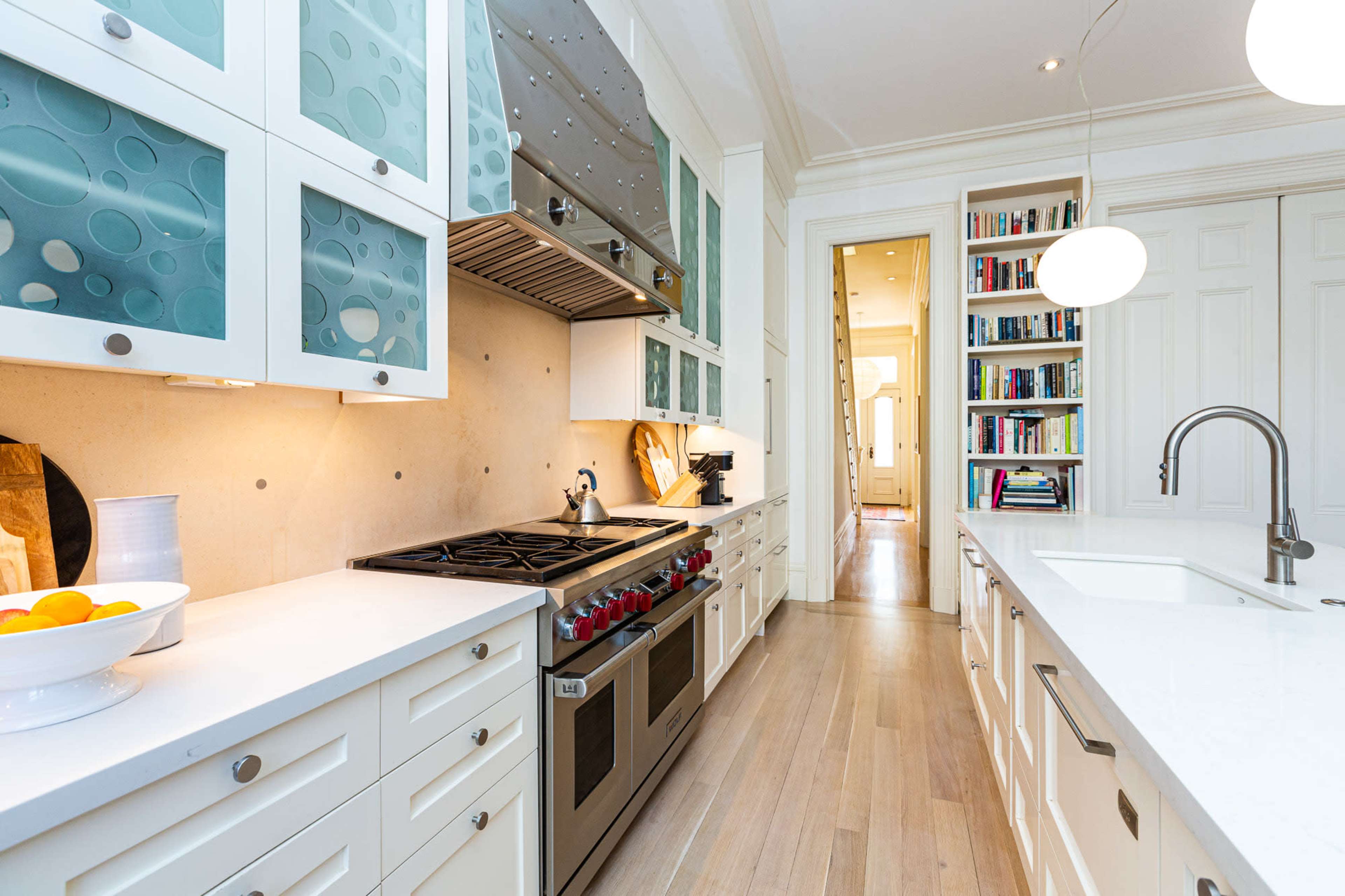 The image shows a modern kitchen with white cabinets, a stainless steel oven, and a long countertop, leading to an adjacent hallway lined with bookshelves.