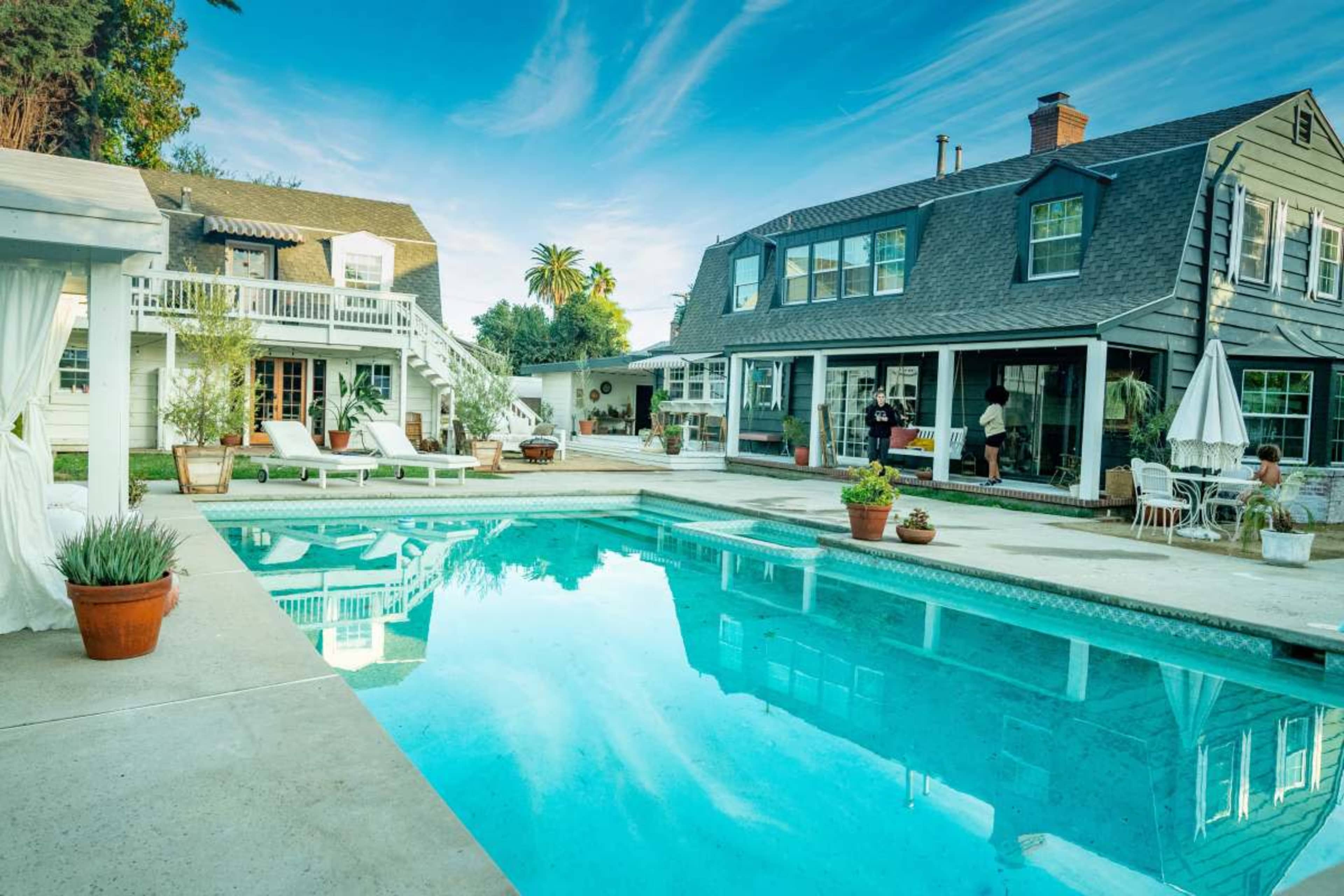 The image shows a spacious backyard with a swimming pool, surrounded by lounge chairs and potted plants, featuring a two-story house and a single-story structure nearby.