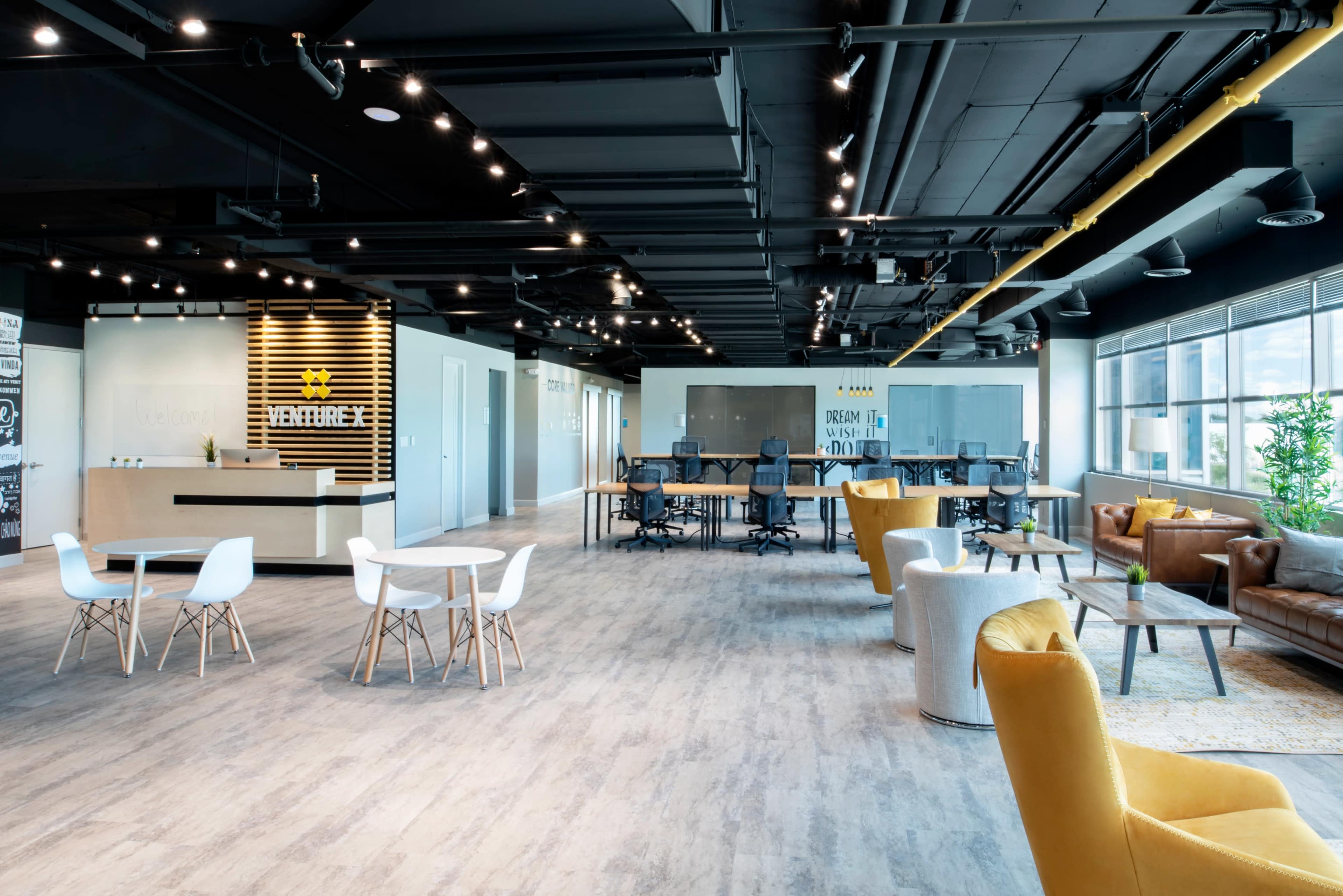 A modern open office space featuring a reception area, multiple seating arrangements, and workstations with large windows allowing natural light.