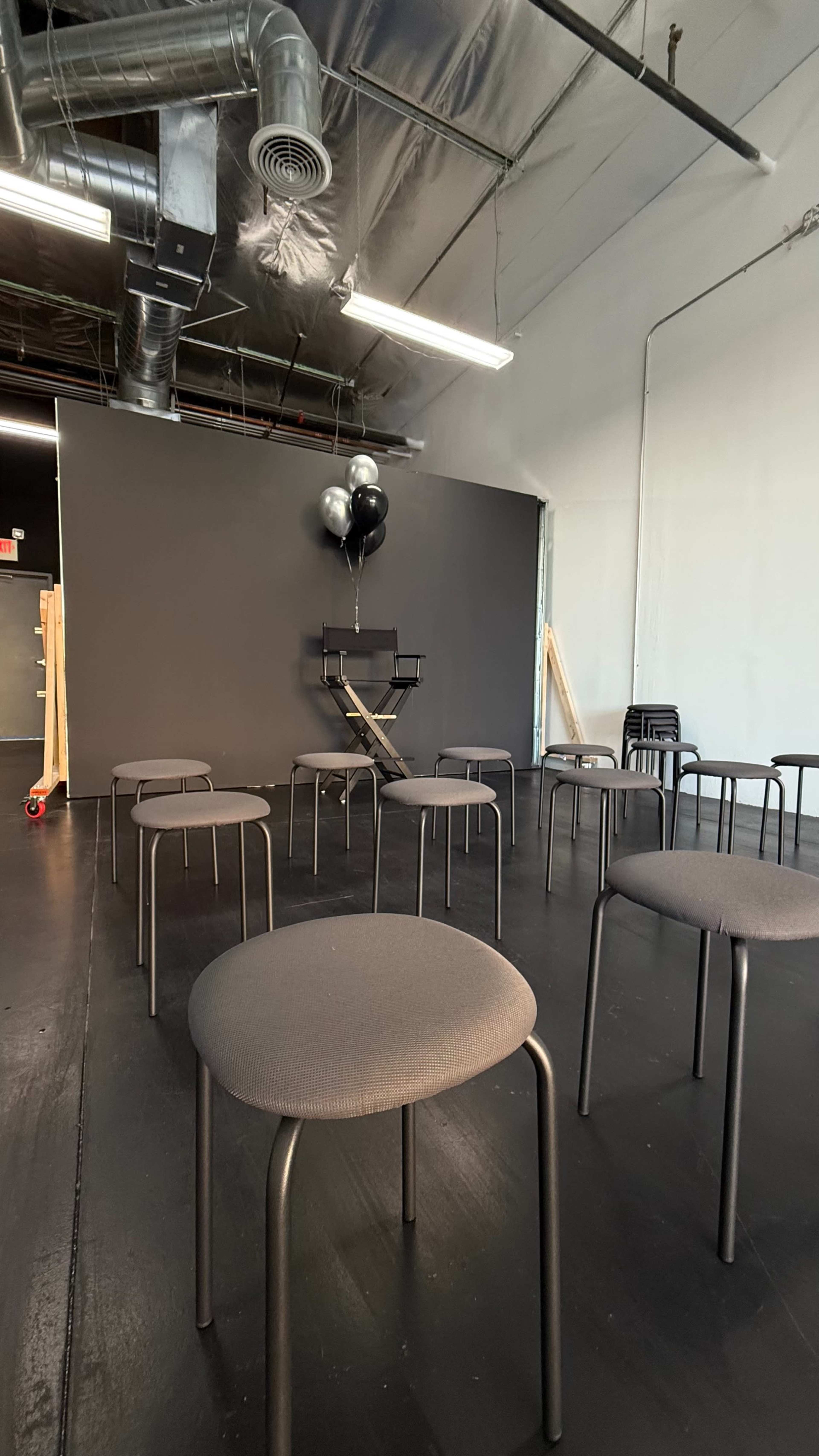 The image shows a room with a dark gray wall, several black chairs arranged in a circular formation, and a small stage with balloons in the background.
