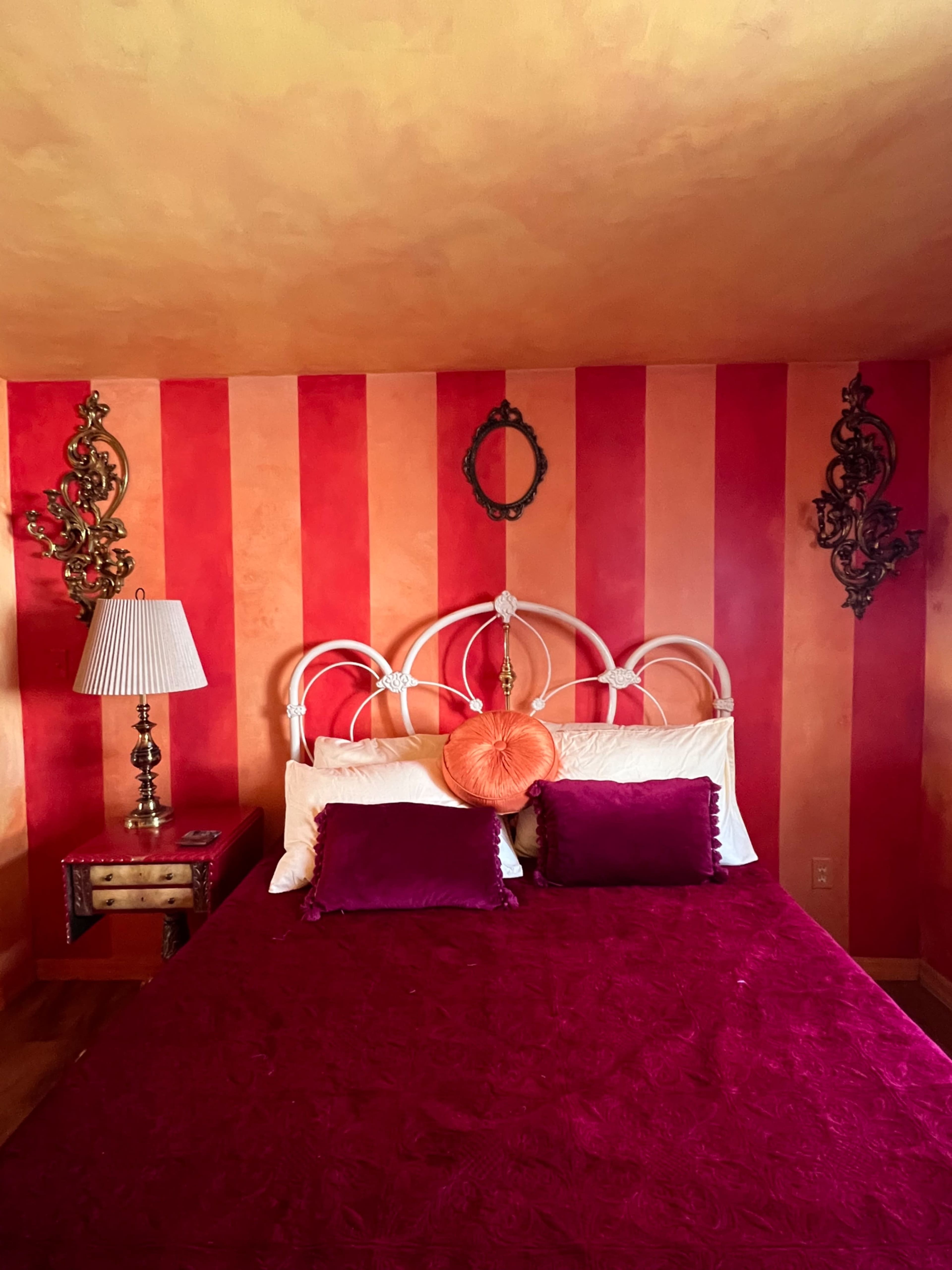The bedroom features a bed with a burgundy quilt, accented by purple pillows, and is set against striped orange and red walls with ornate wall decorations and a bedside lamp.