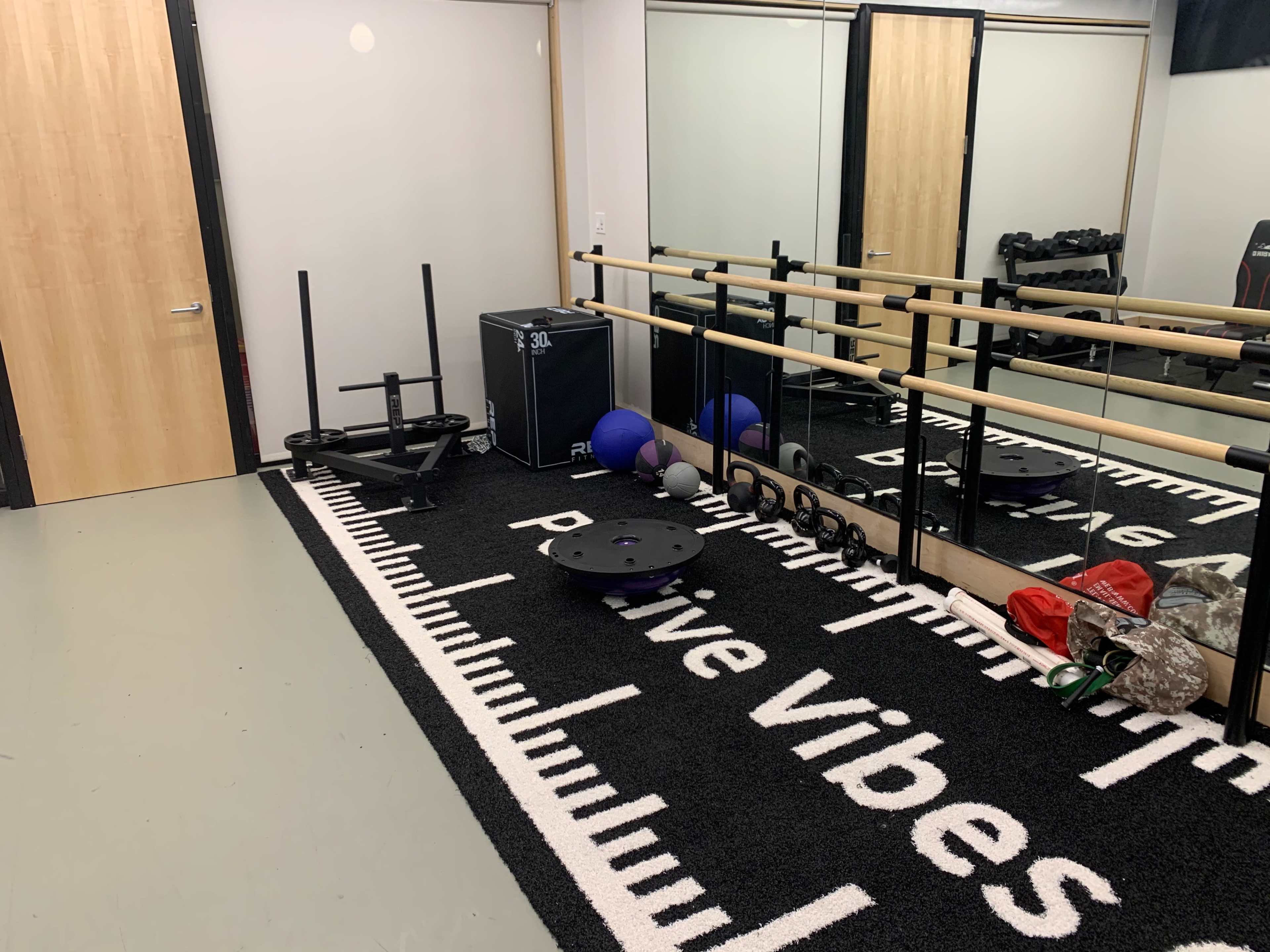 The image shows a small gym area with a black and white rug, exercise equipment including weights, a sled, and a mirror along one wall.