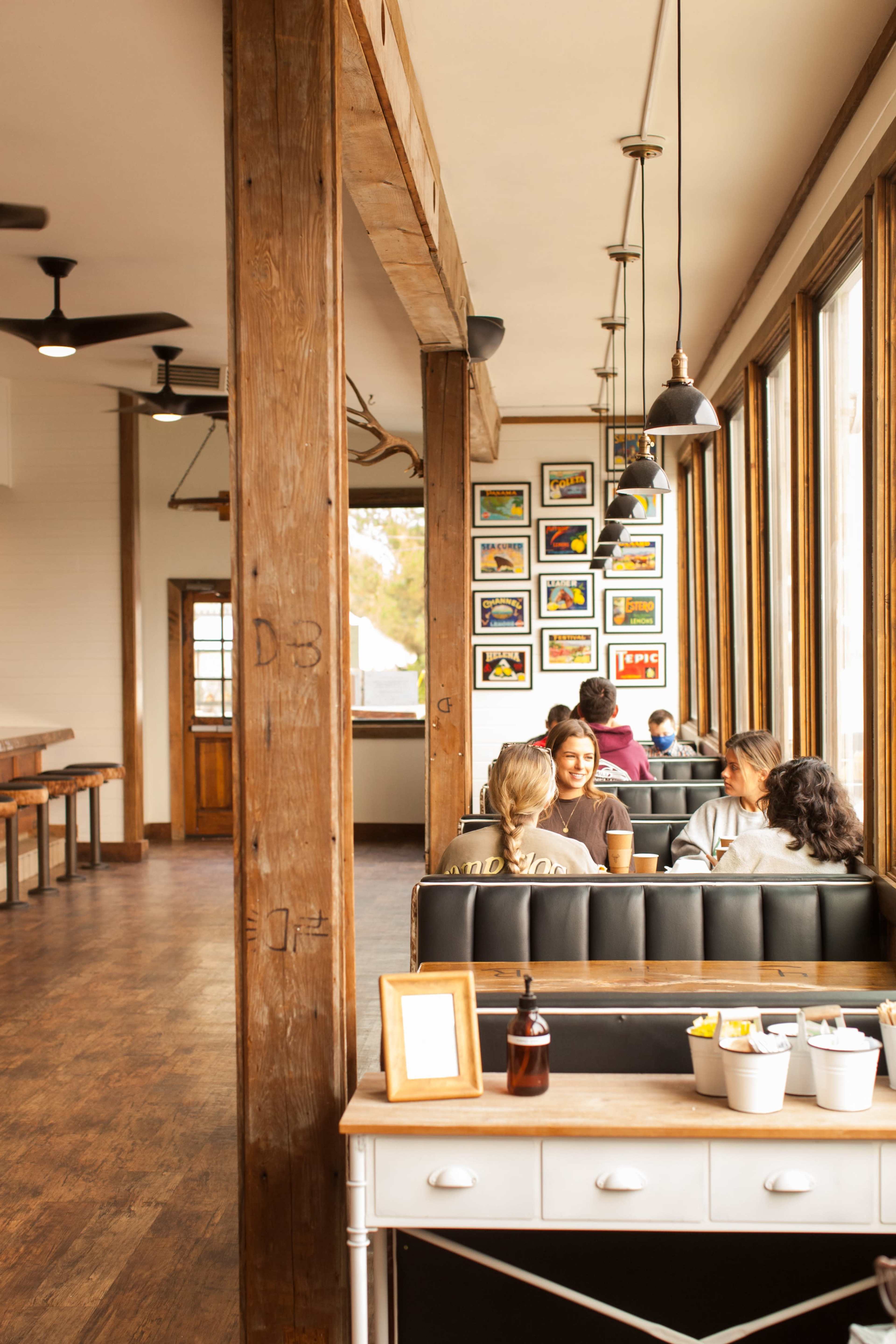 A modern café interior with wooden beams, large windows, and customers seated at booths, engaged in conversation.