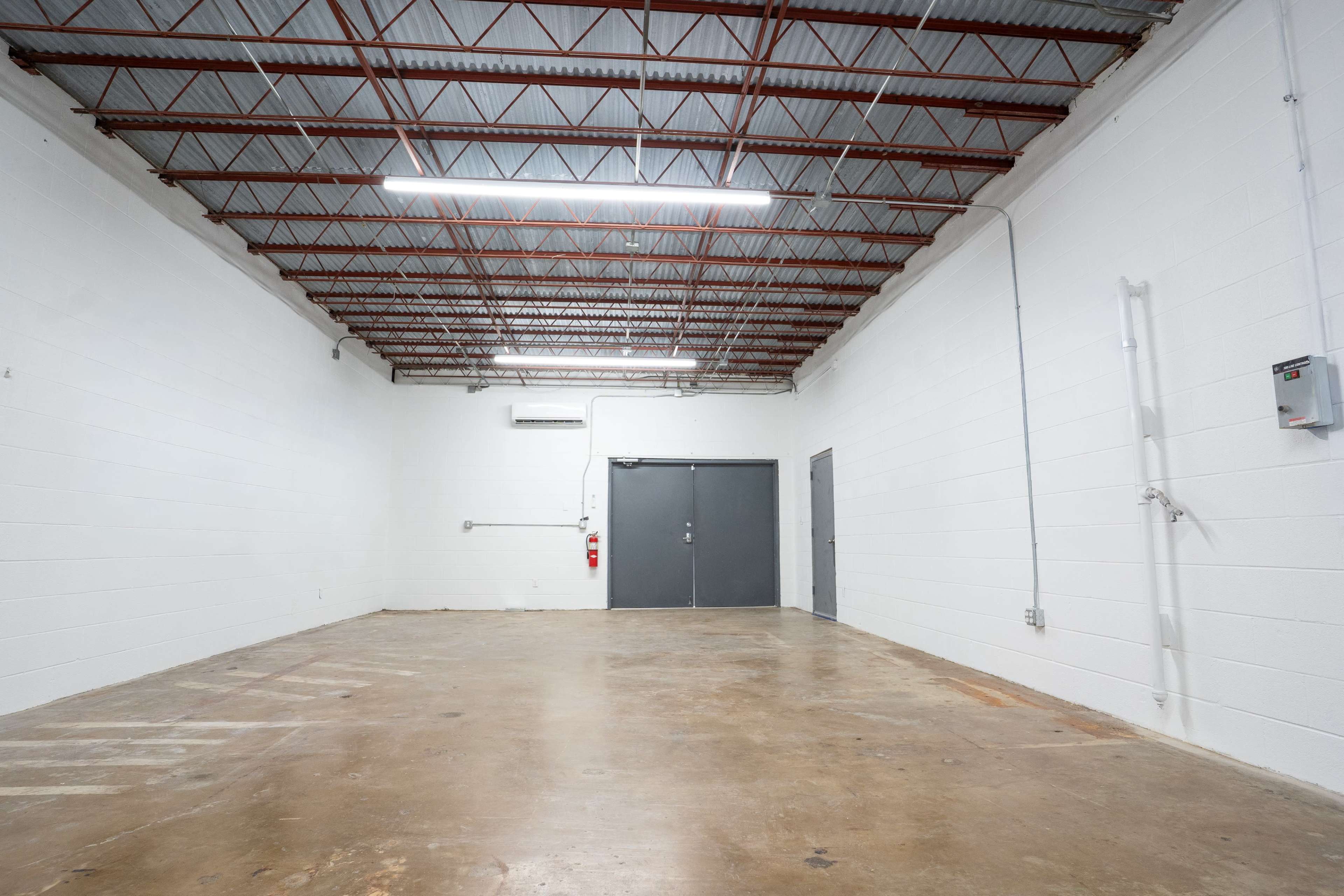 An empty warehouse space with a concrete floor, white walls, and a metal ceiling, featuring two gray doors and minimal electrical fittings.