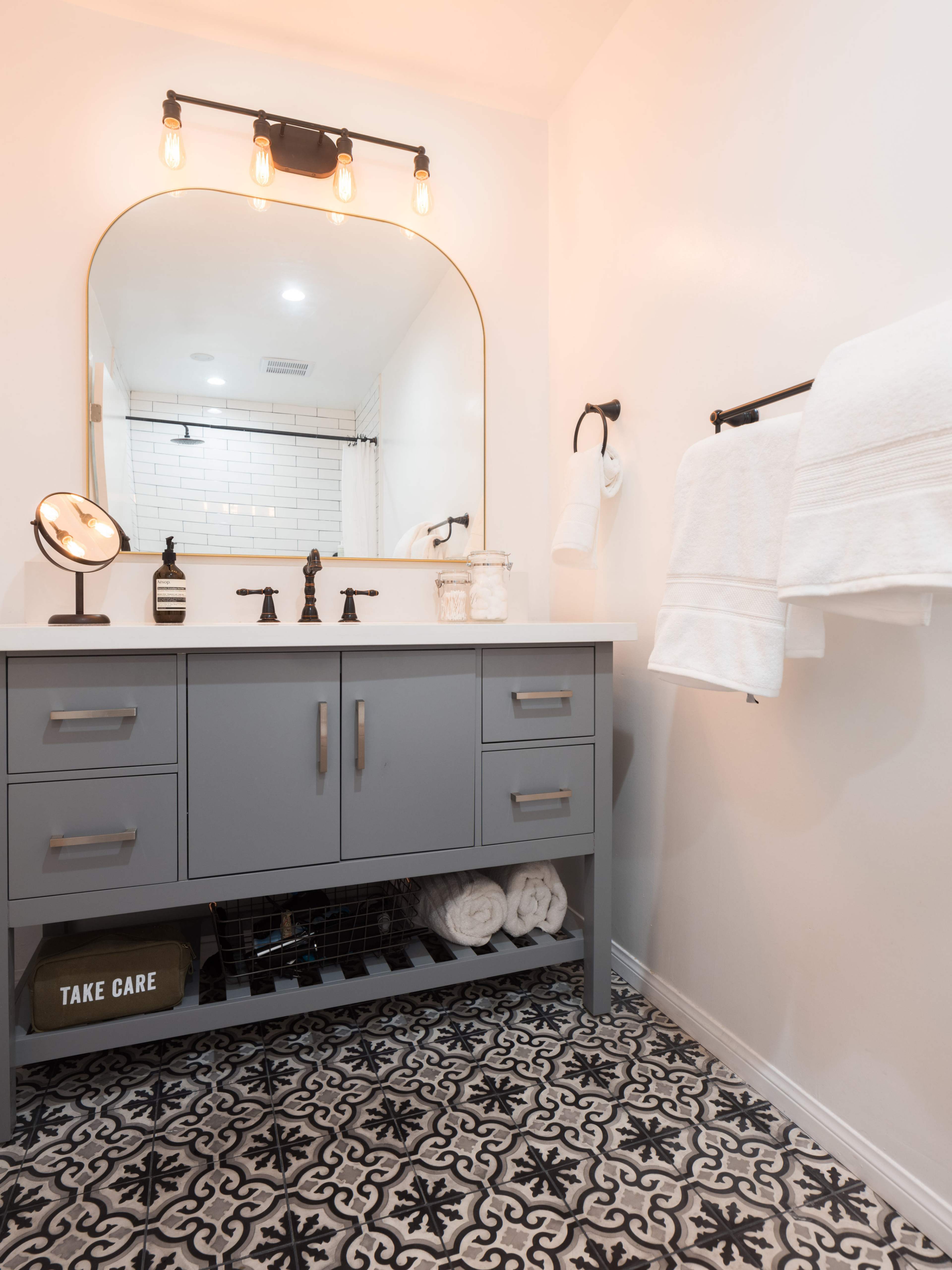 The image shows a modern bathroom with a gray vanity, a large mirror, and patterned floor tiles.