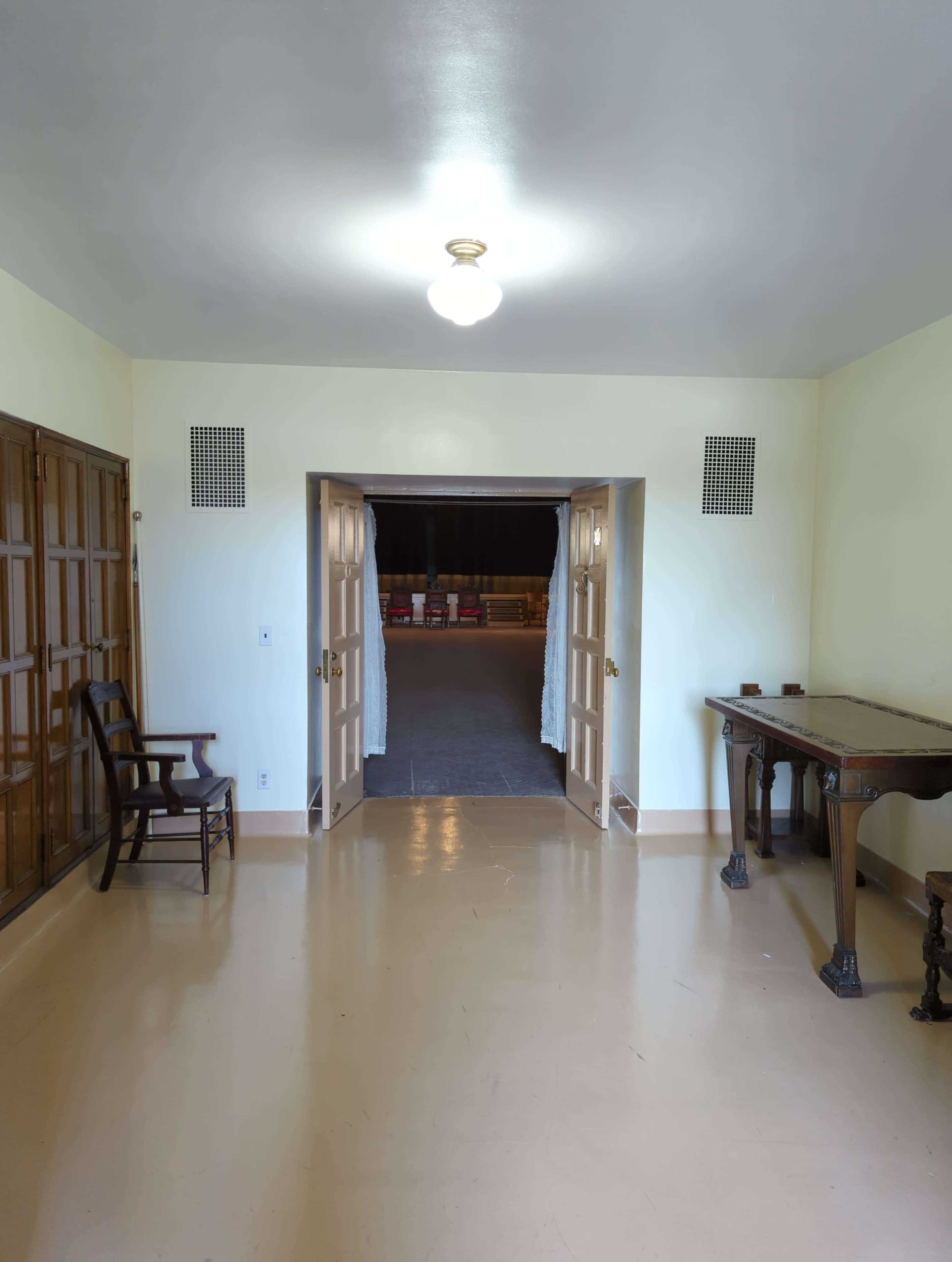 A brightly lit room with a wooden chair and table, featuring an open doorway leading to a dimly lit area.