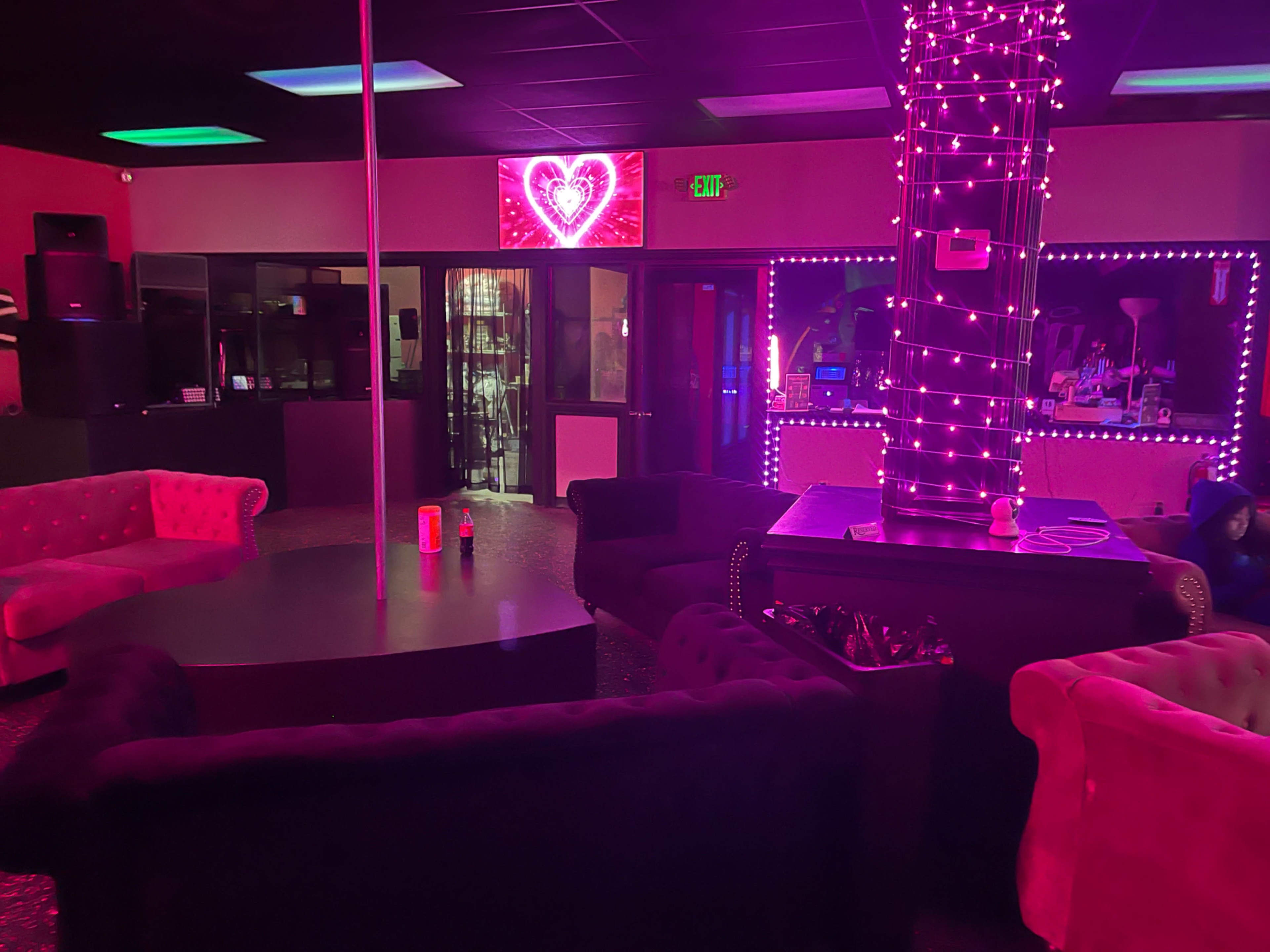 A dimly lit lounge with pink and purple lighting, featuring plush seating arranged around a central pole and illuminated decorative elements.