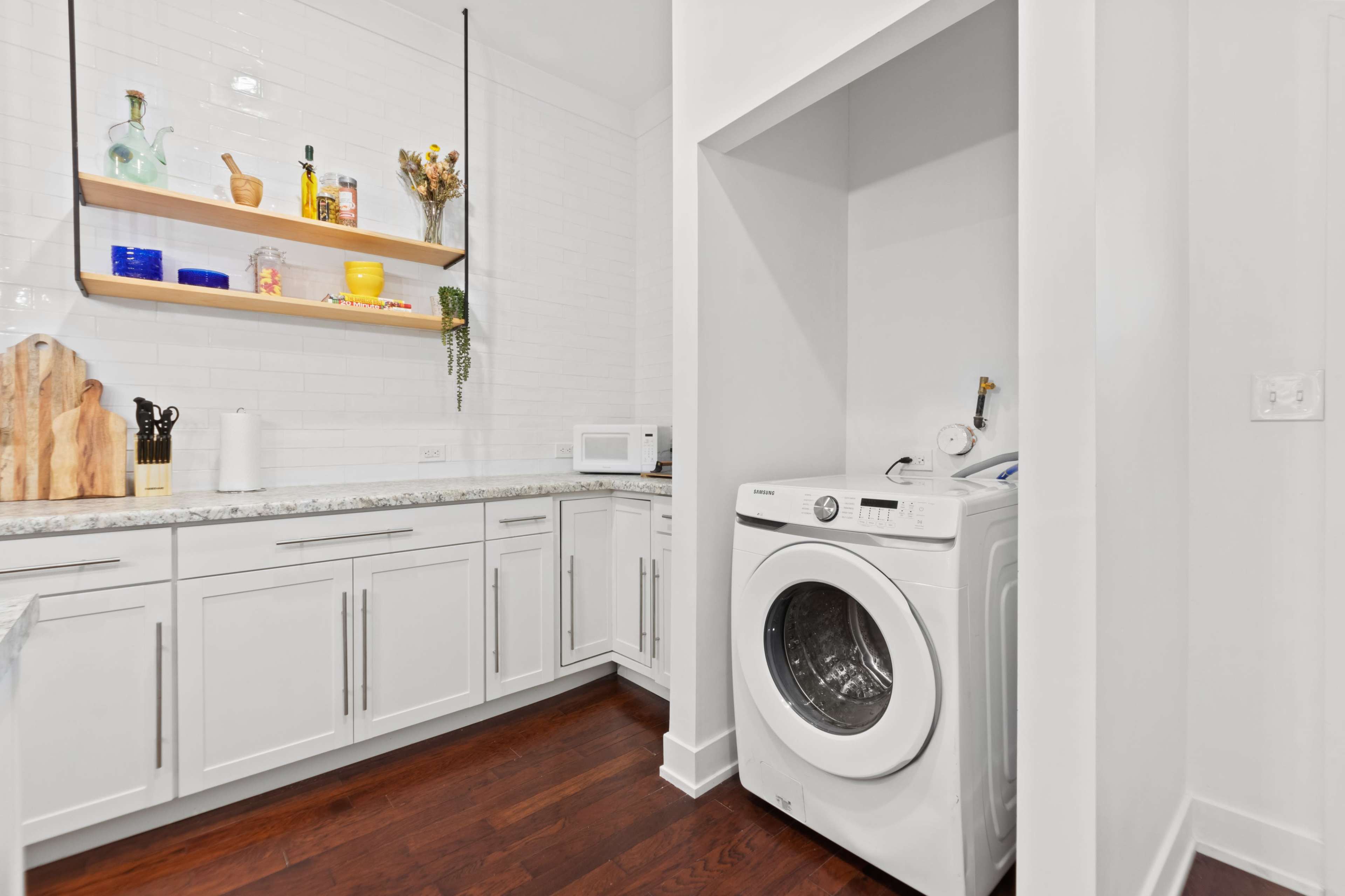 A laundry room featuring a washer, a countertop, shelves with various items, and a microwave.