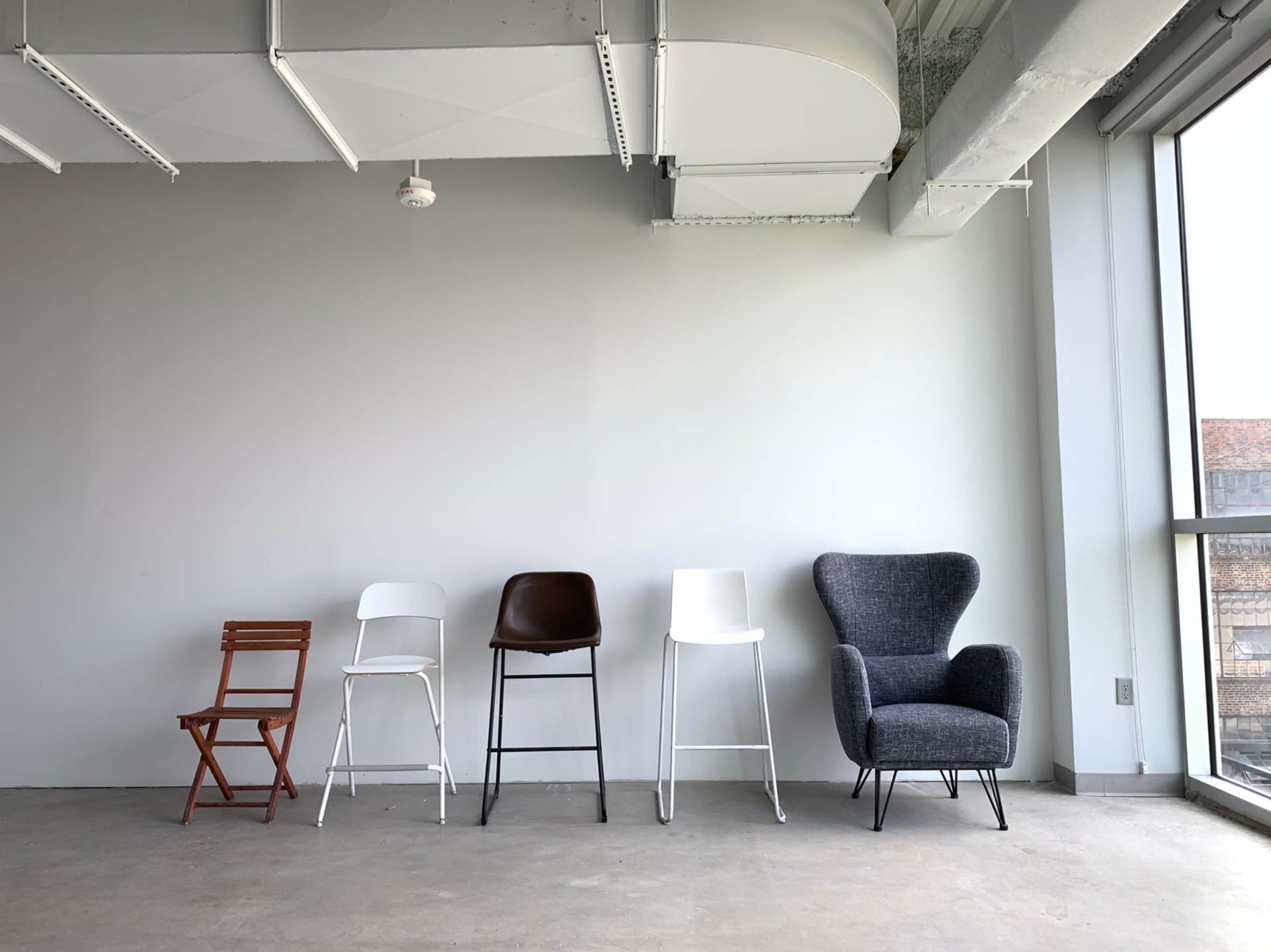 A row of five different chairs placed against a plain gray wall in a well-lit interior space.