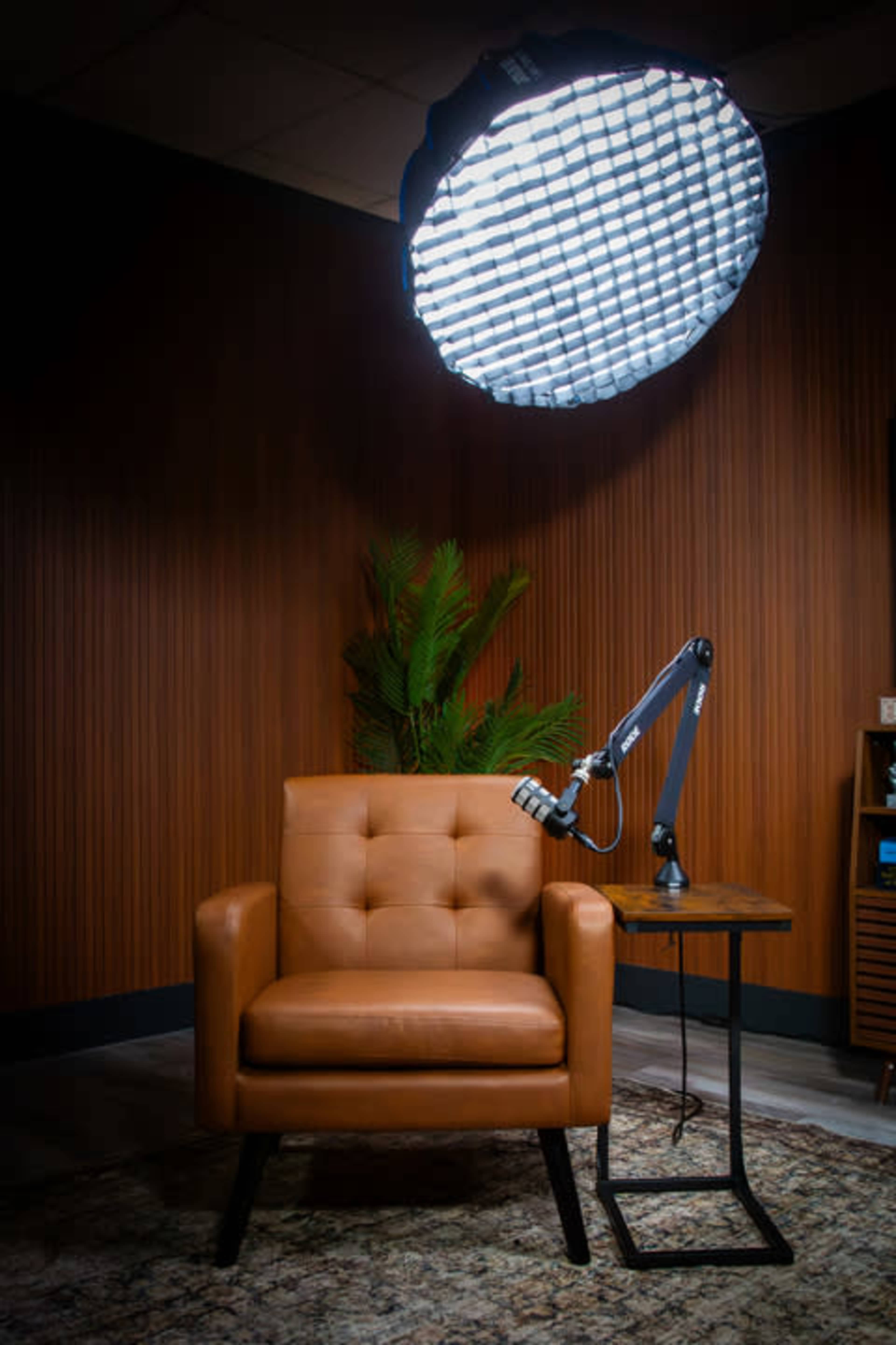 The image shows a brown upholstered armchair next to a small round side table, with a large overhead light and a plant in the background.