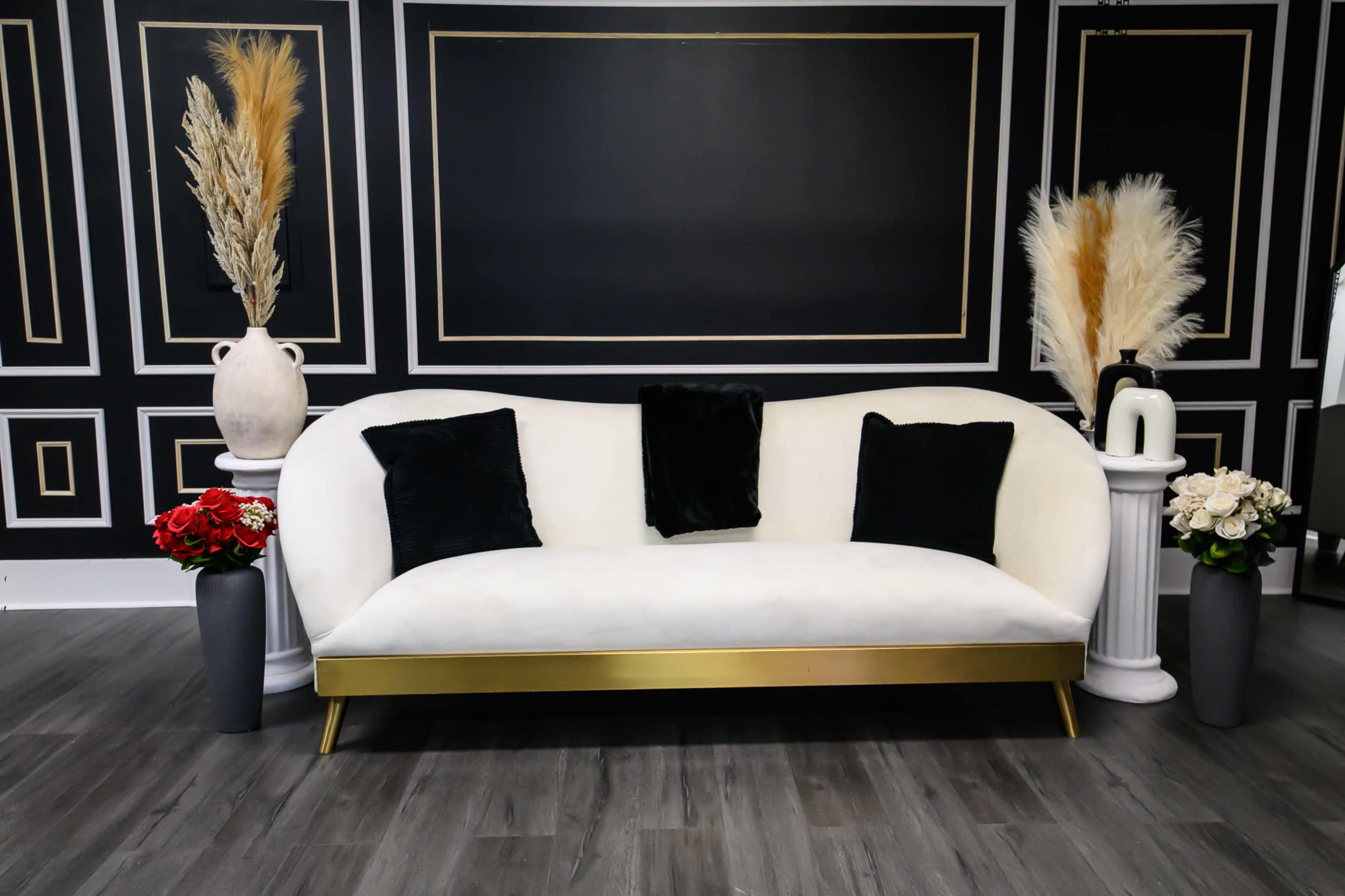 A modern white sofa with black pillows is positioned against a black wall adorned with gold trim, flanked by vases of flowers and decorative plants.