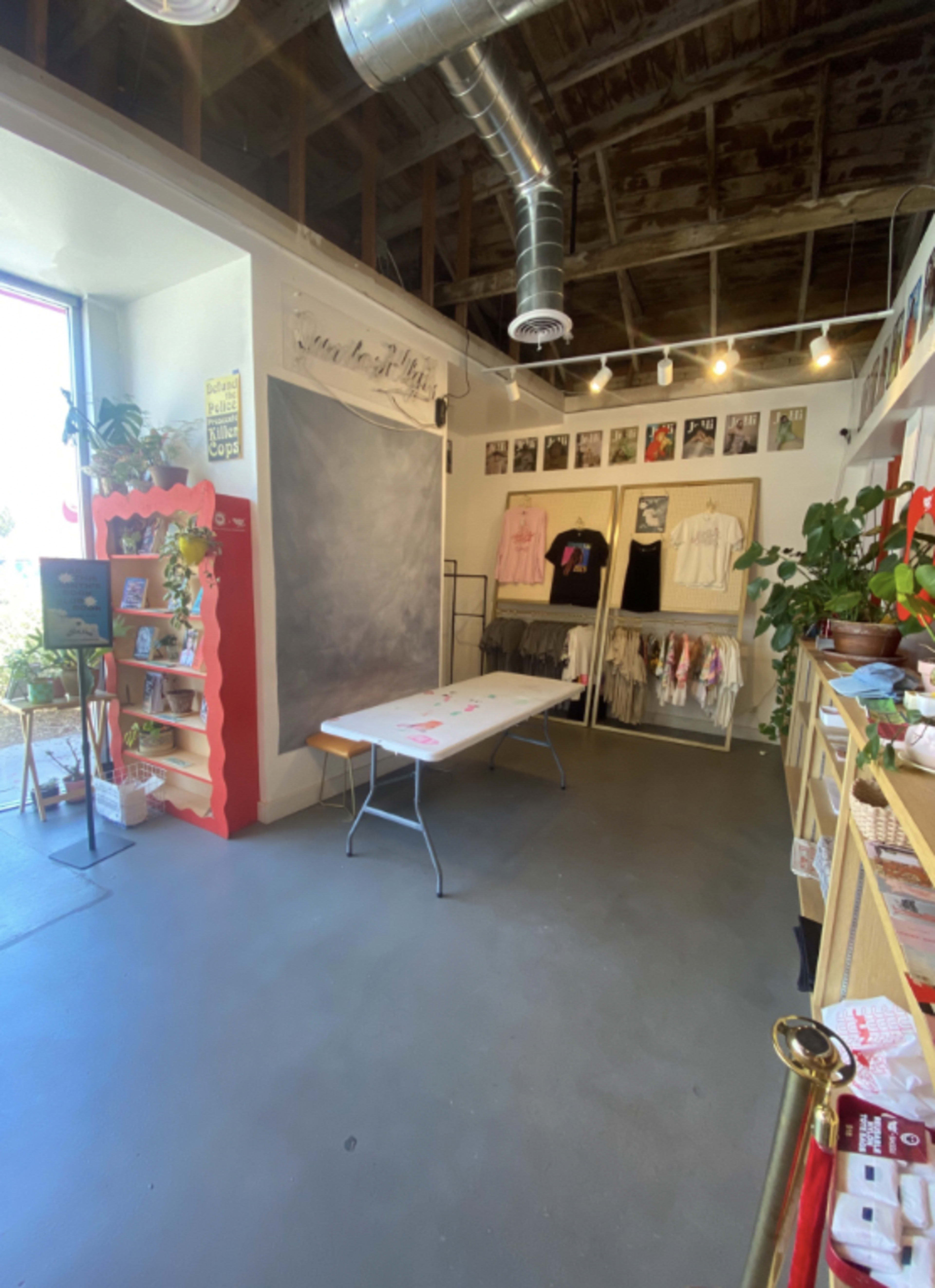 The interior of a small retail space with clothing racks, a folding table, plants, and a large chalkboard wall.