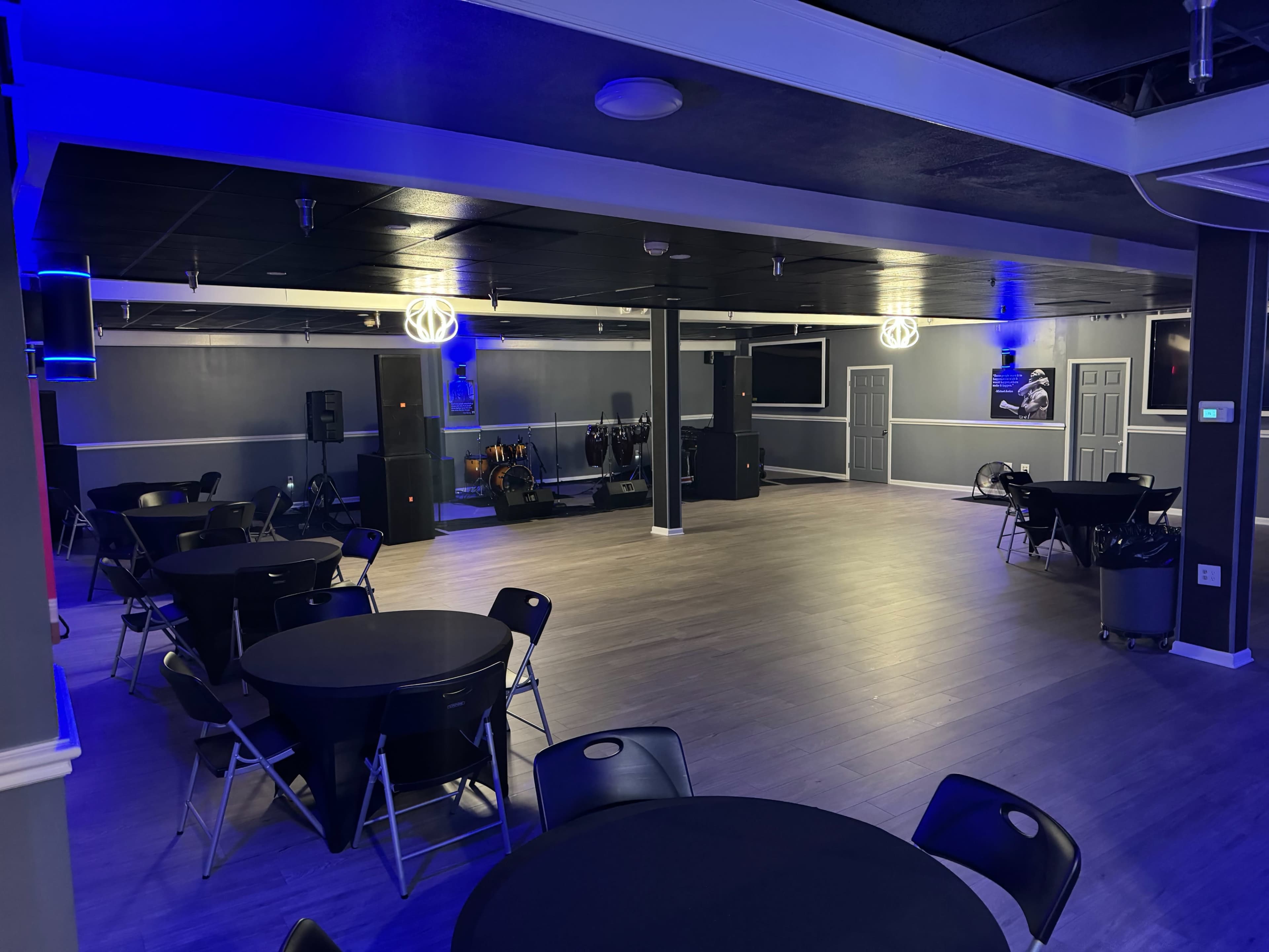 A spacious event room features black tables and chairs, with a stage area and blue lighting along the walls.
