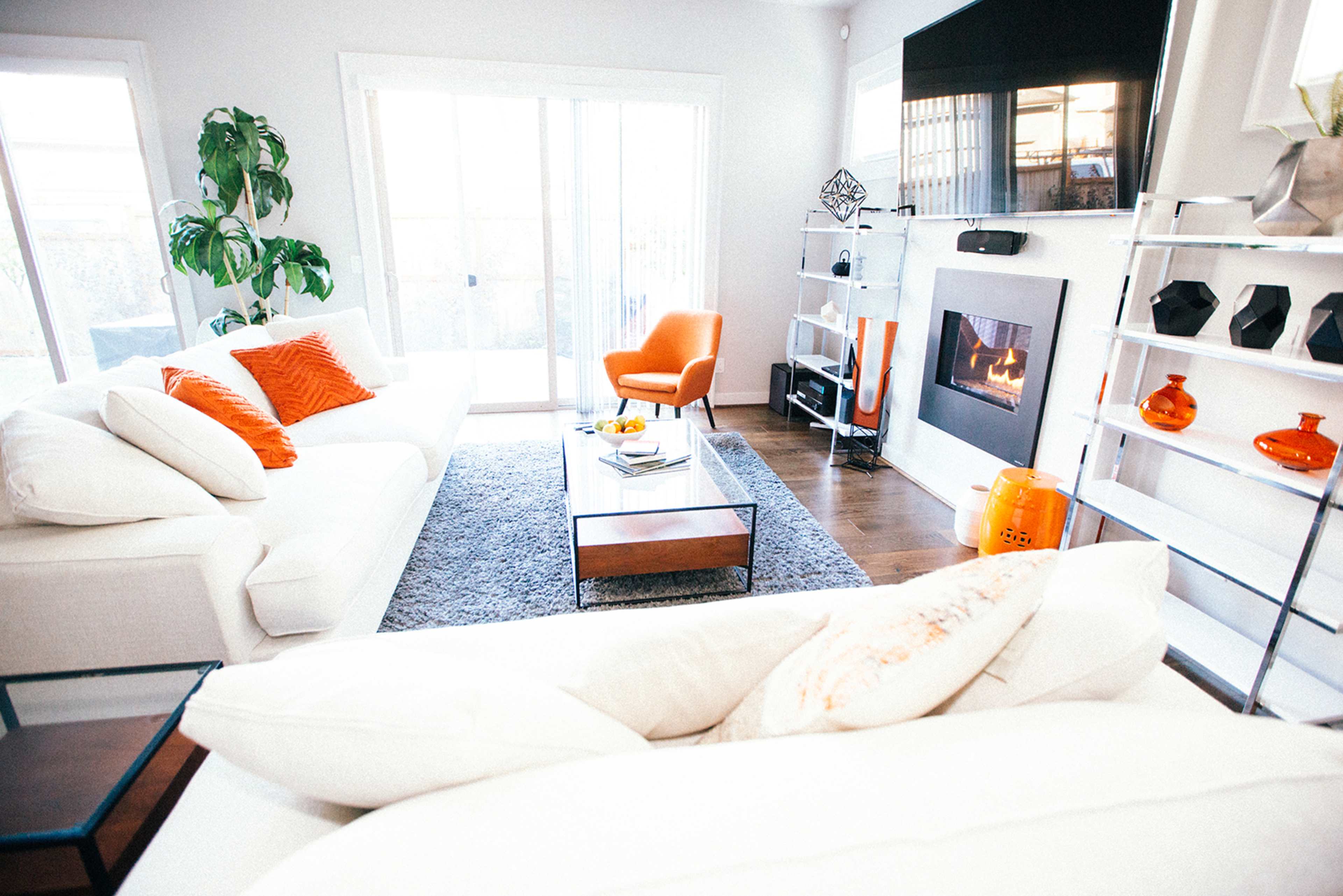 A bright living room features white sofas, an orange armchair, a wooden coffee table, and a fireplace with a contemporary design.