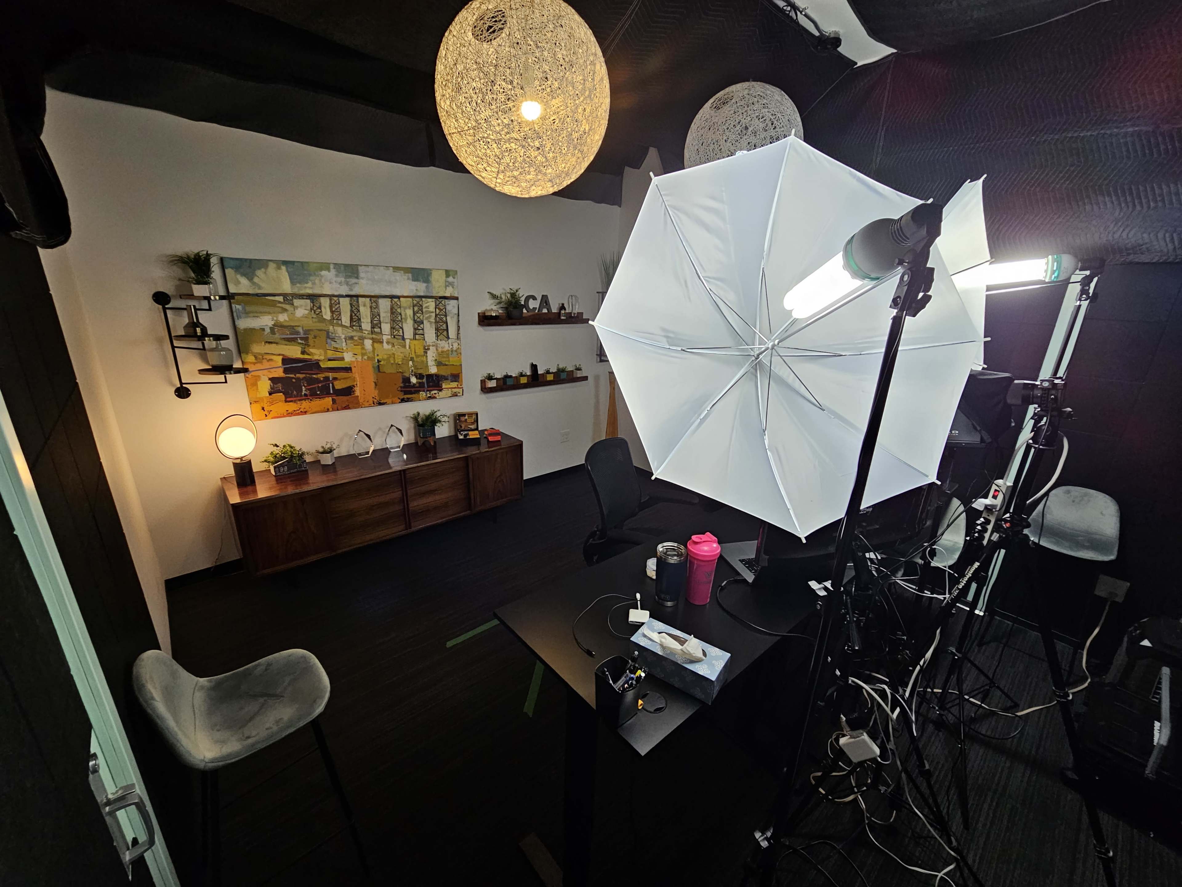 A small studio space with a wooden cabinet, a black desk, lighting equipment, and decorative elements arranged in a modern setting.