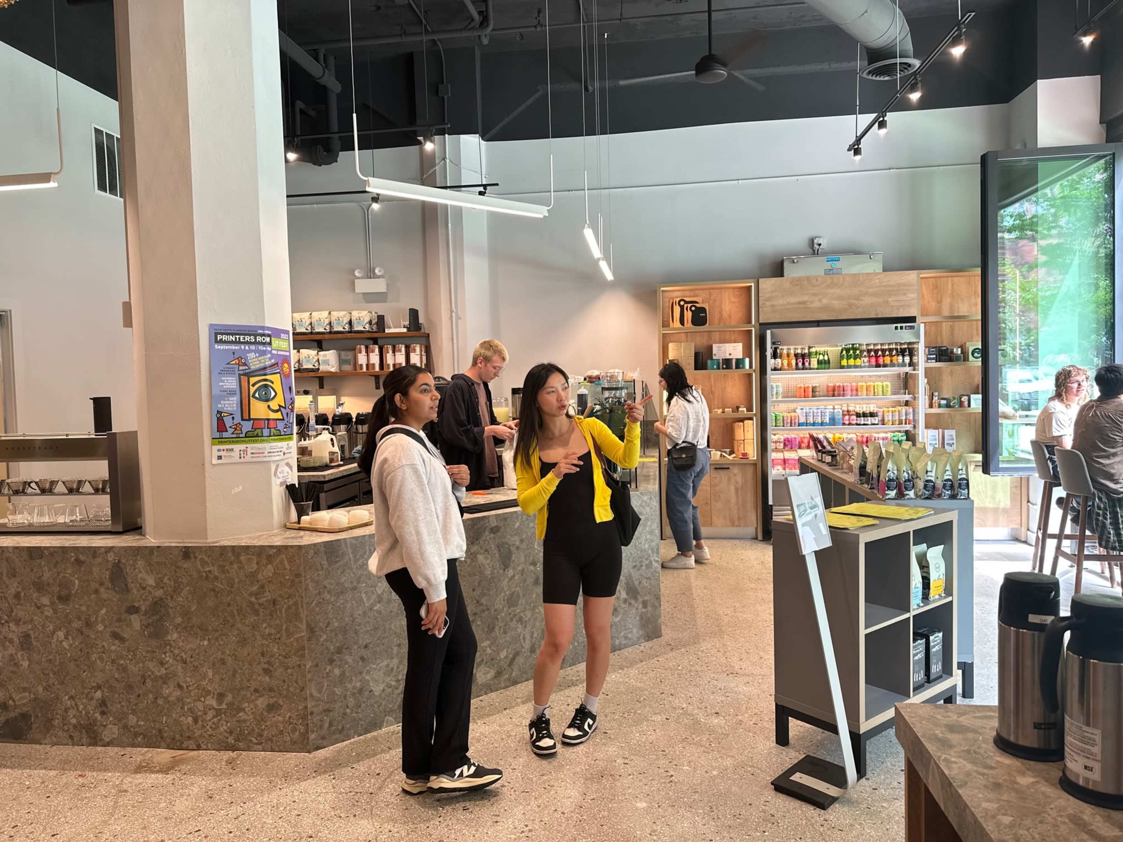 Two women converse in a coffee shop featuring a large counter, shelves of drinks, and a few patrons seated at tables.
