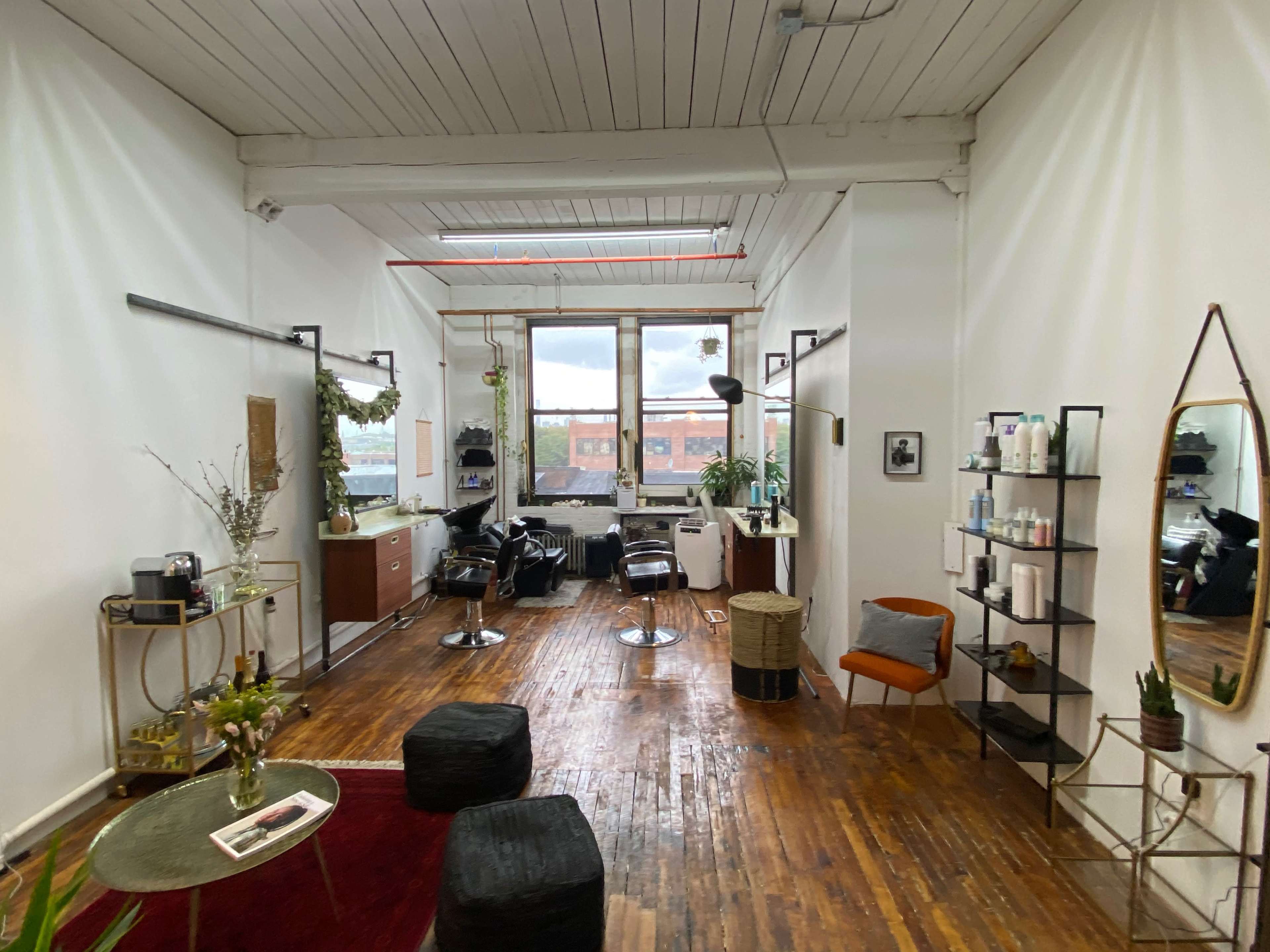 A well-lit hair salon with wooden floors, a large window, and several styling stations equipped with chairs and mirrors.