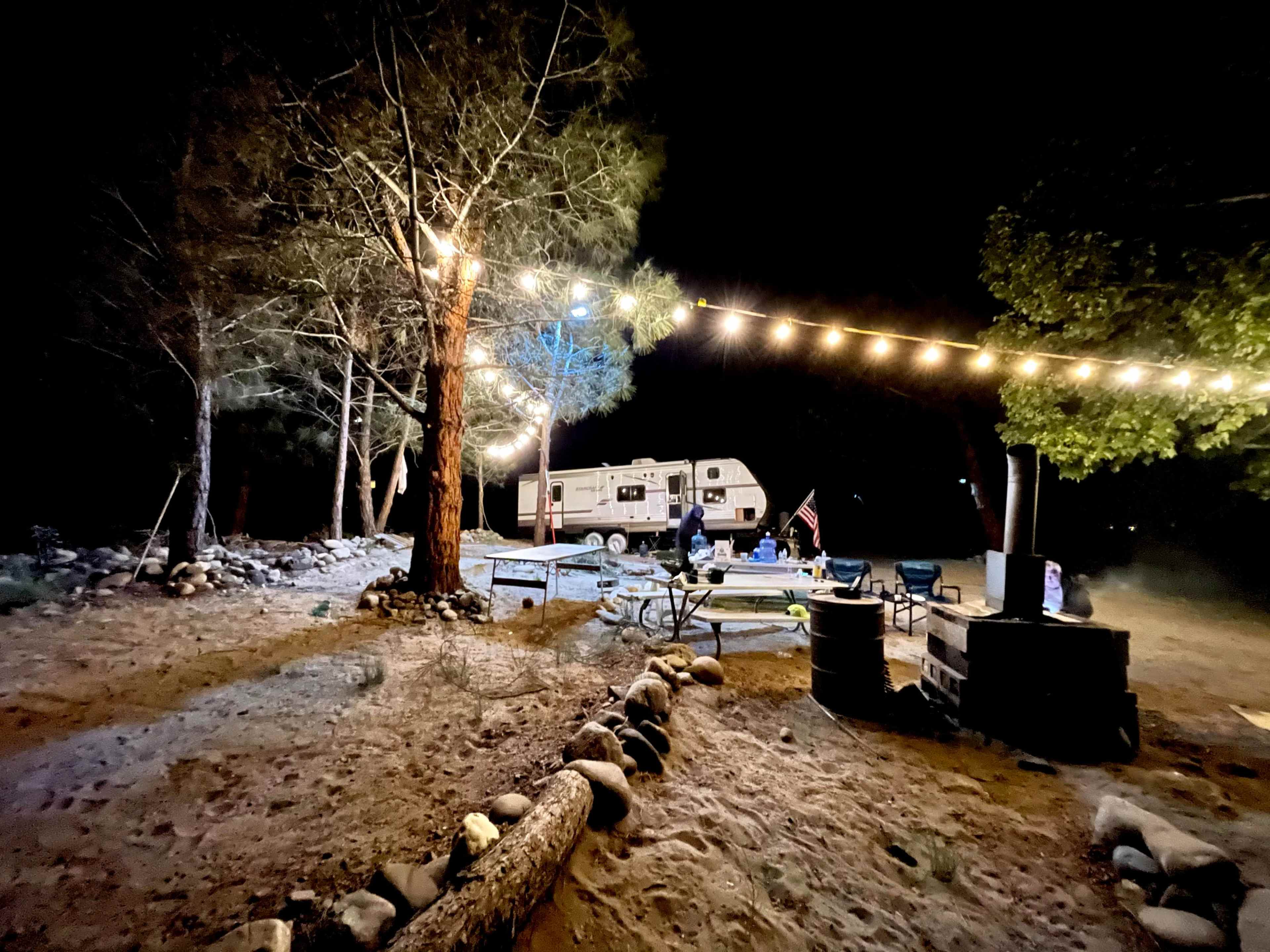 A campsite at night features a parked RV, string lights illuminating a picnic area, and a campfire surrounded by seating.