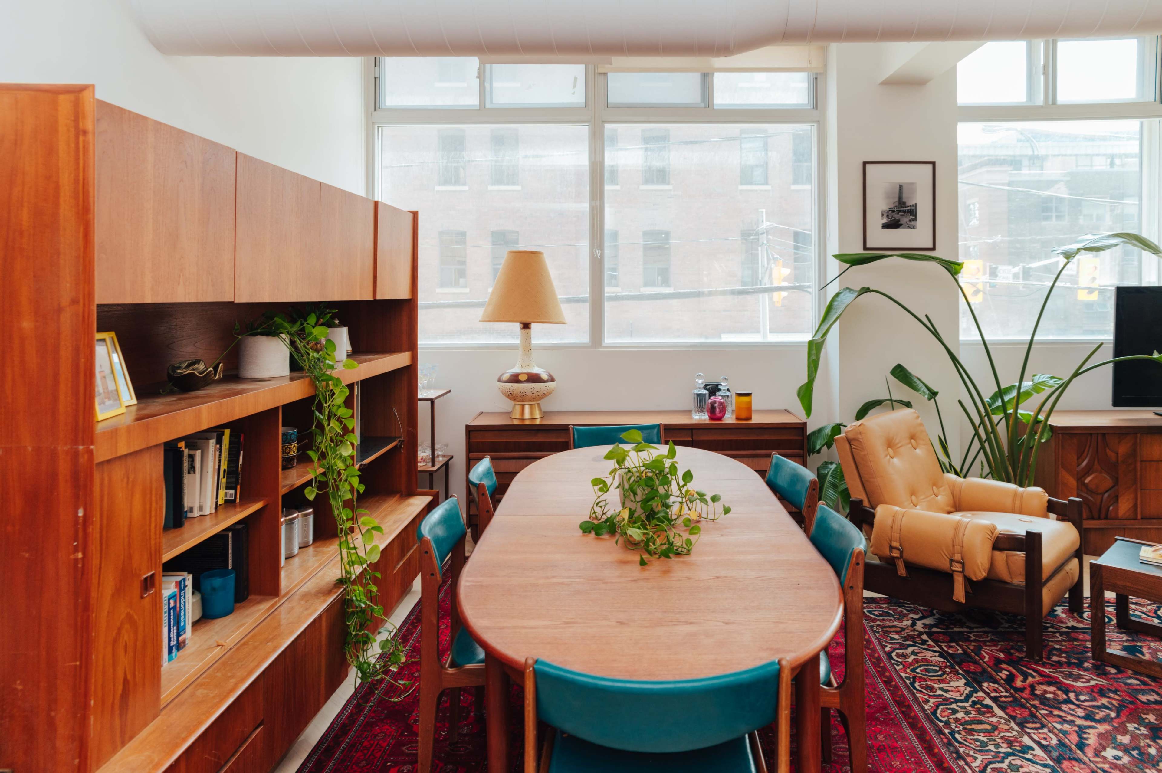 The image shows a modern workspace with a wooden dining table surrounded by teal chairs, shelves filled with books, and large windows letting in natural light.