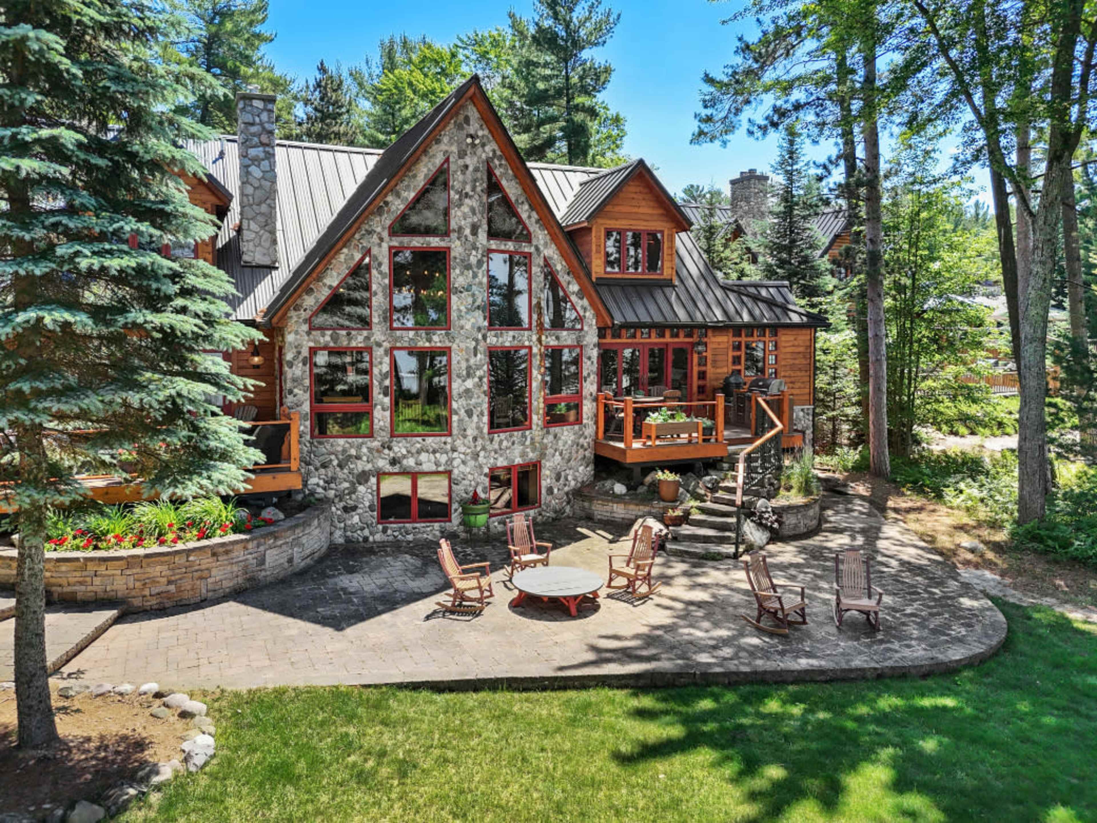 A large, modern cabin with stone and wood exteriors features multiple windows and a circular seating area on a stone patio surrounded by trees.