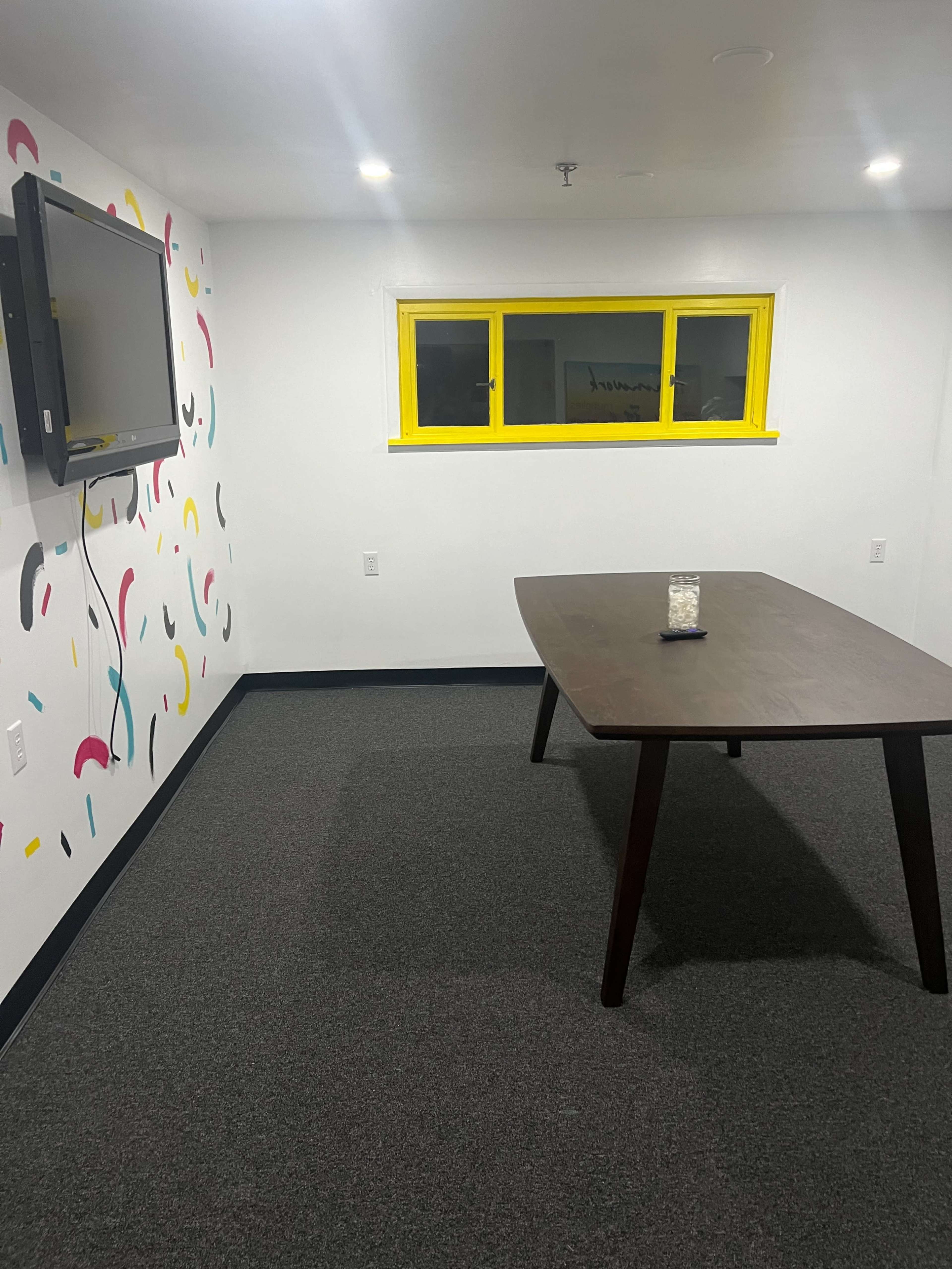 A brightly lit meeting room features a large table, a television mounted on the wall, and a window with yellow trim.