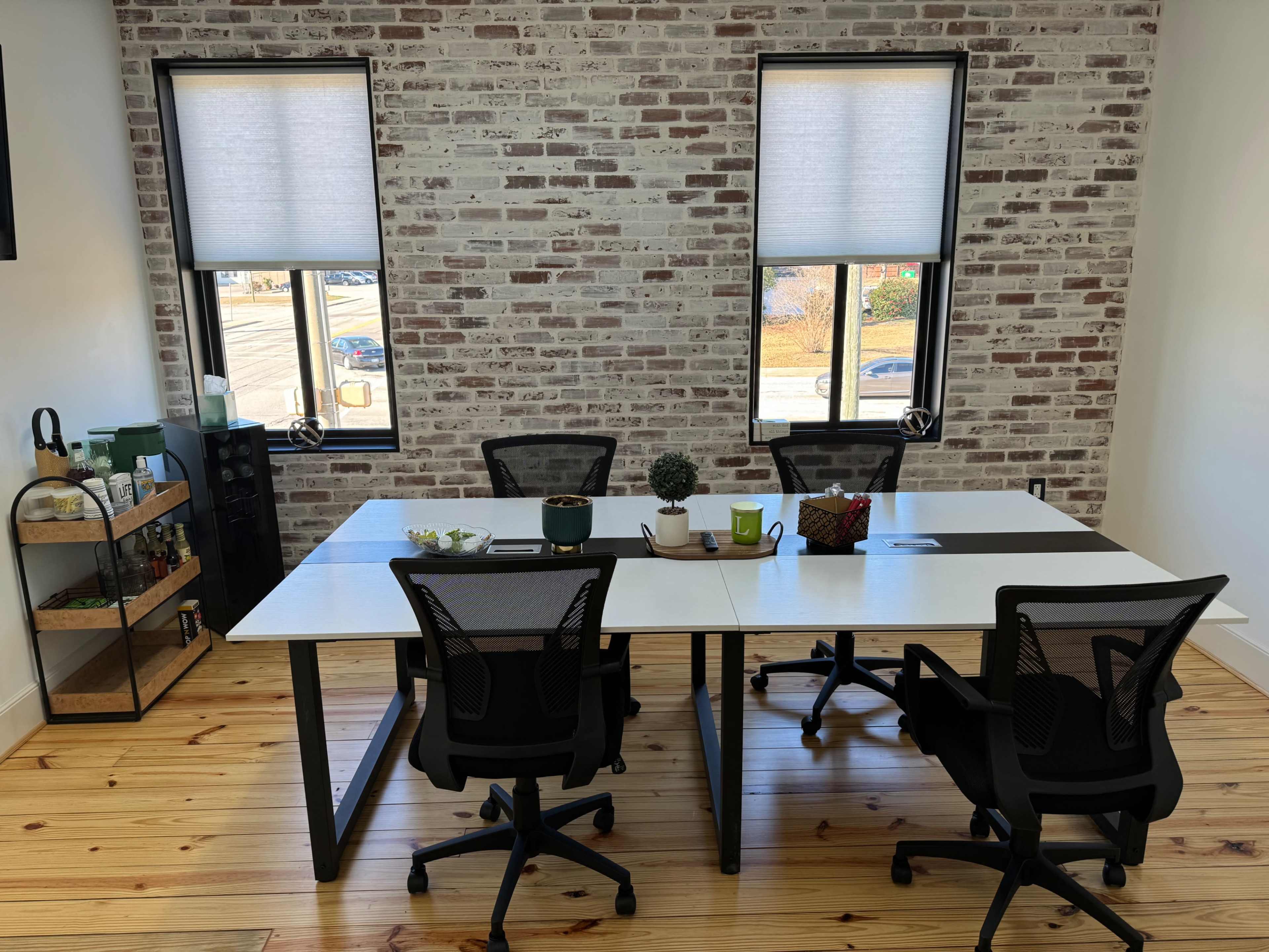 A modern conference room features a large table surrounded by black office chairs and two windows with blinds, set against a brick wall.