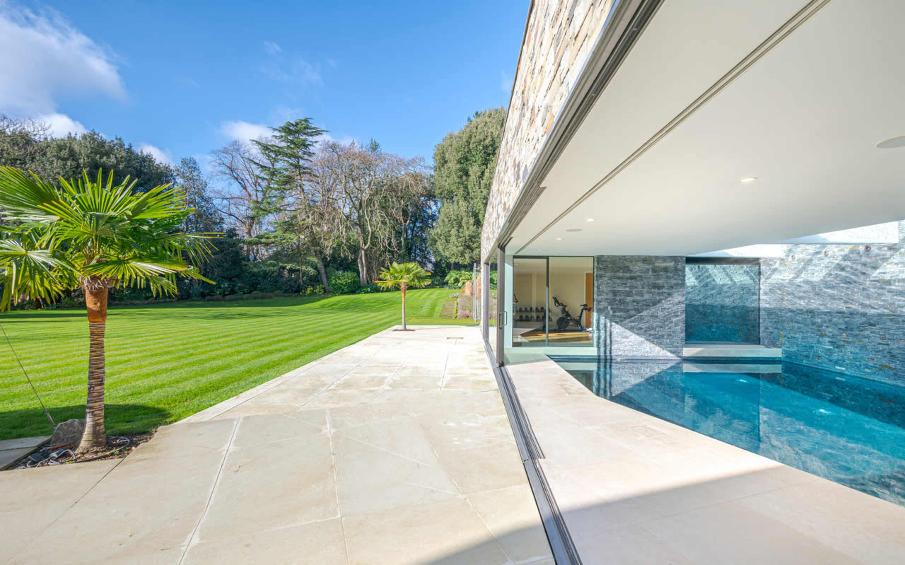 A modern home with a glass wall overlooking a landscaped garden, featuring a swimming pool and a palm tree.