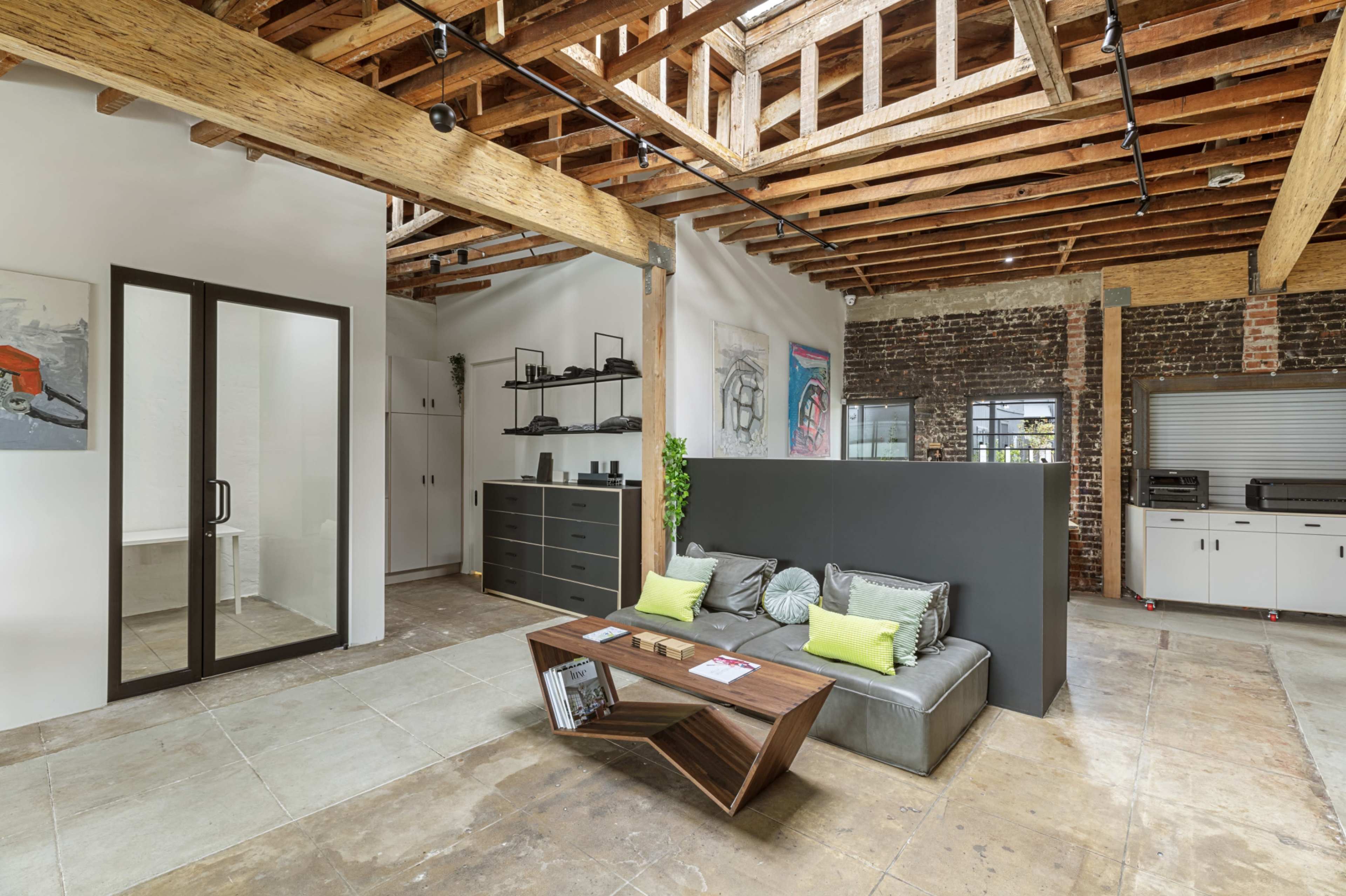 The image shows a modern open-concept office space featuring a sitting area with a gray sofa and a wooden coffee table, exposed wooden beams, and a brick wall with shelves and a bathroom area visible in the background.