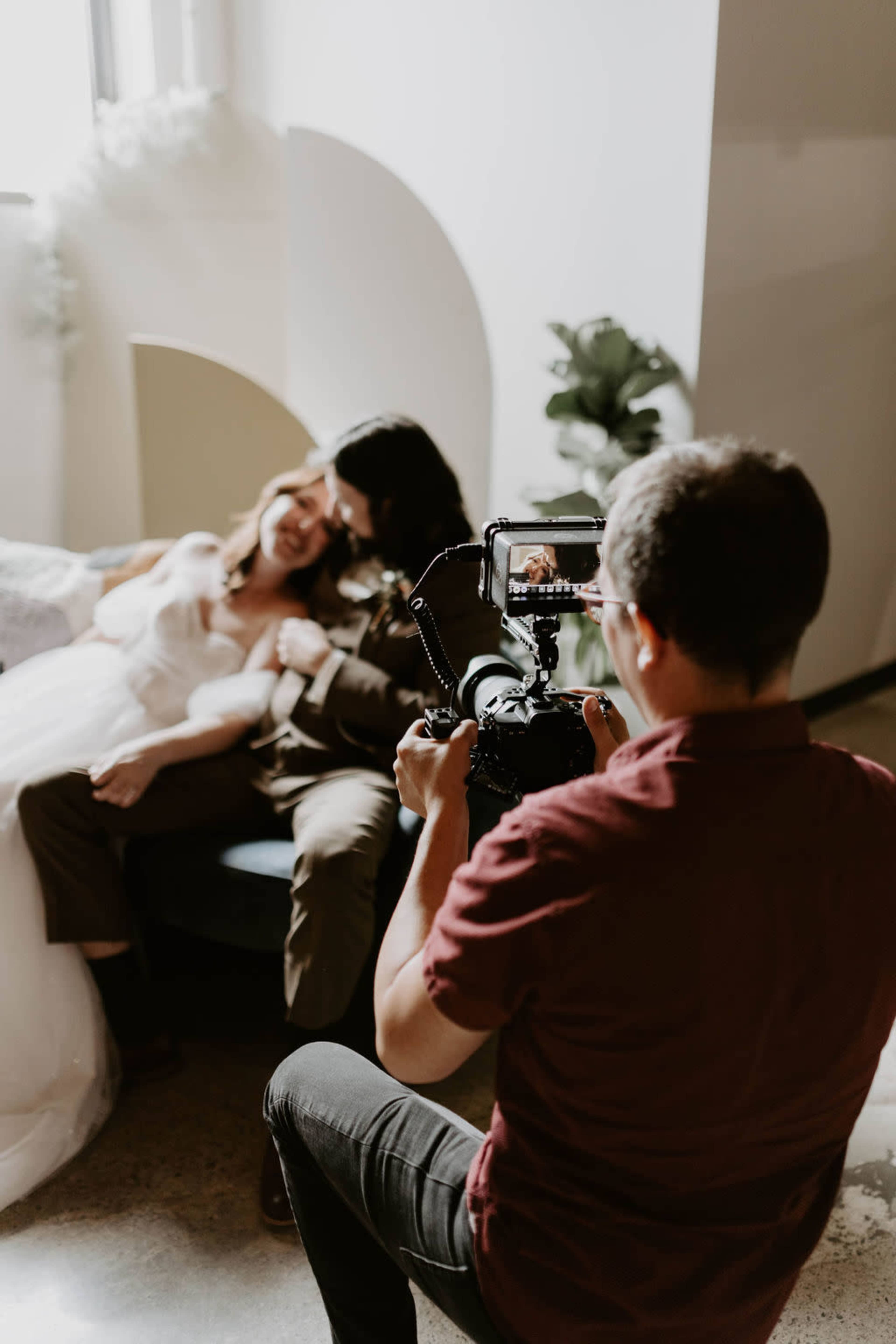 A photographer captures a couple sitting together on a couch in a softly lit interior space.