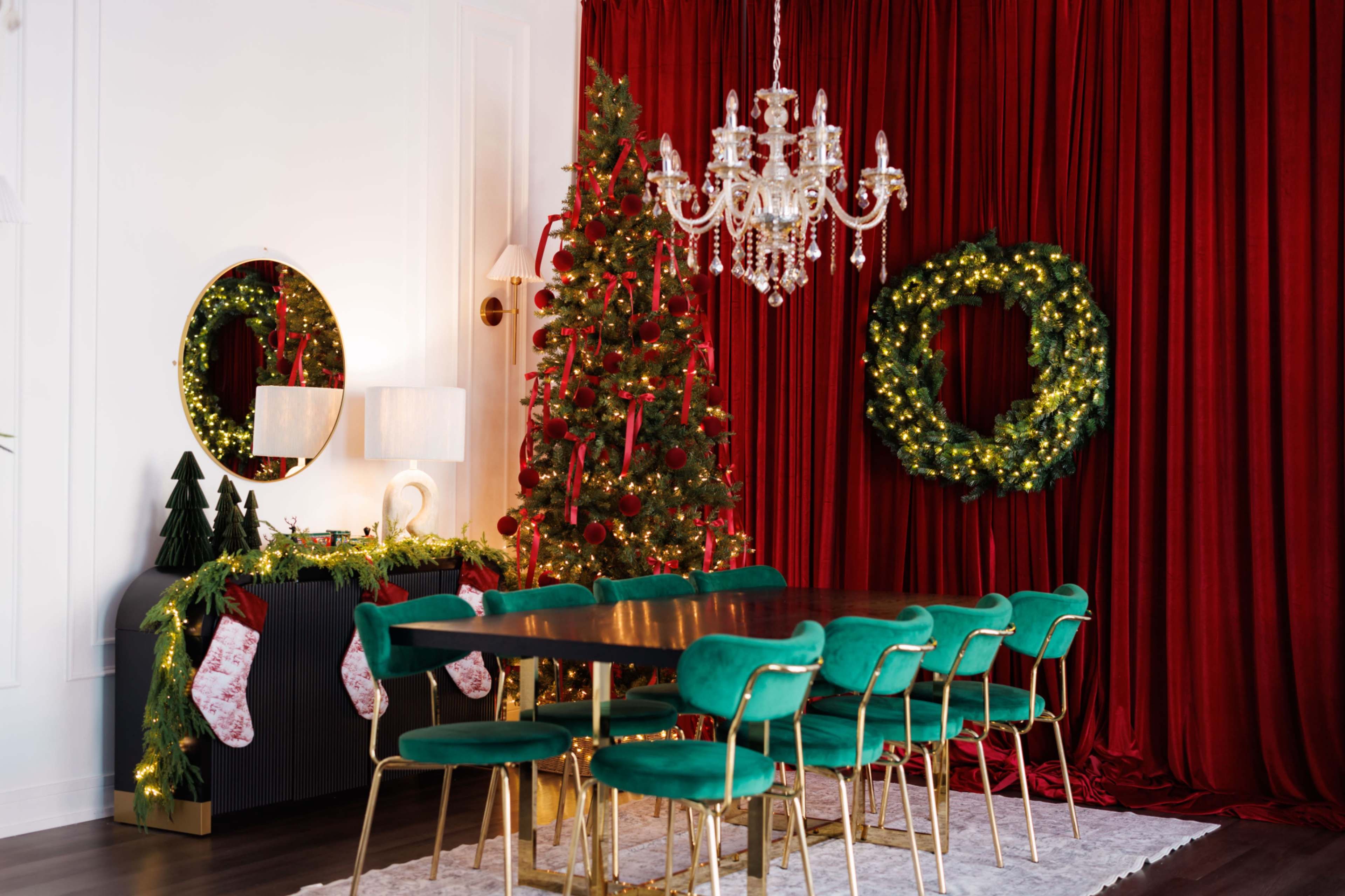 A dining room features a large table surrounded by green chairs, with a decorated Christmas tree and wreaths against a red curtain backdrop.