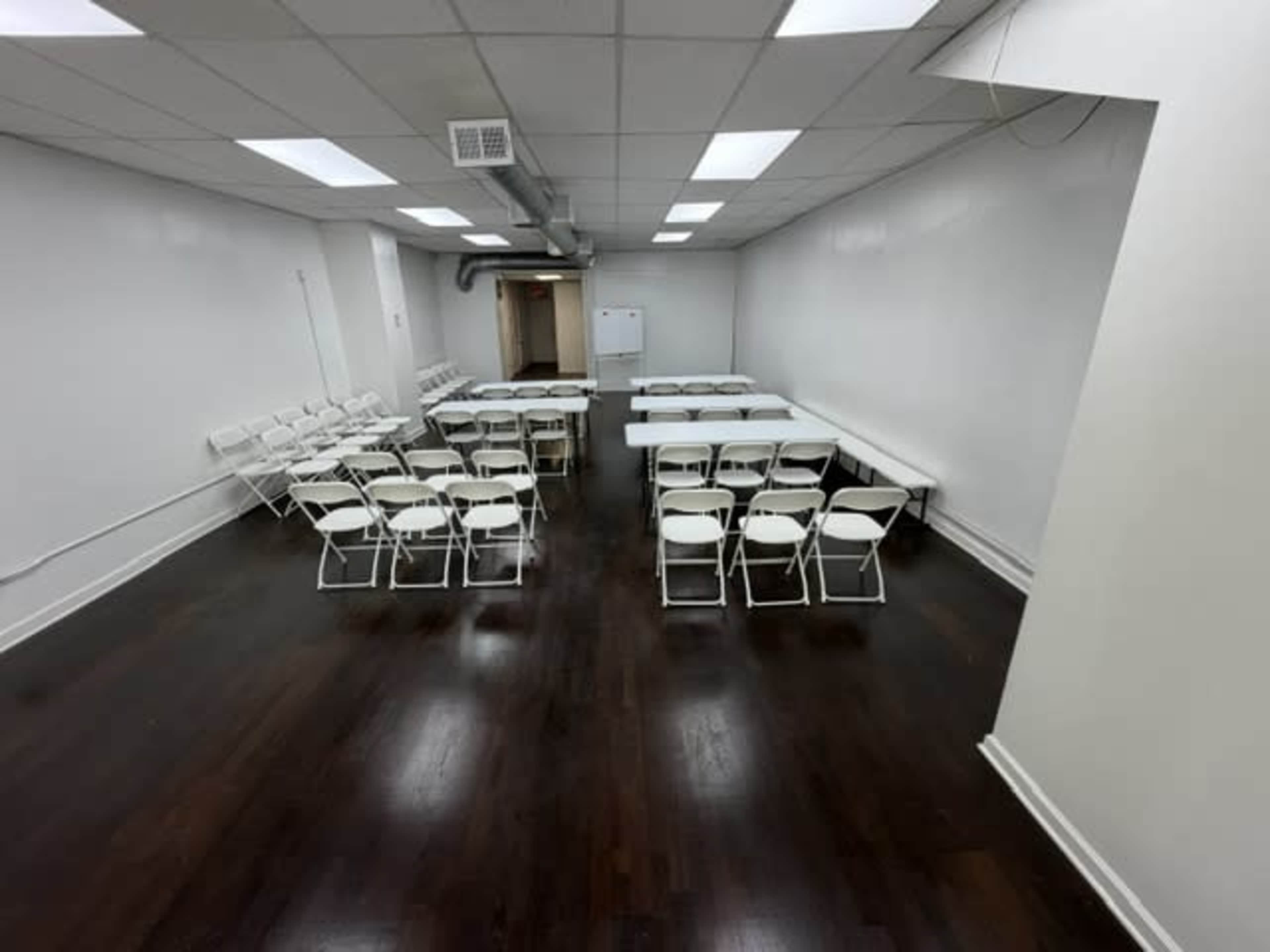 A large, empty room features a setup of white folding chairs arranged around several tables on a hardwood floor.