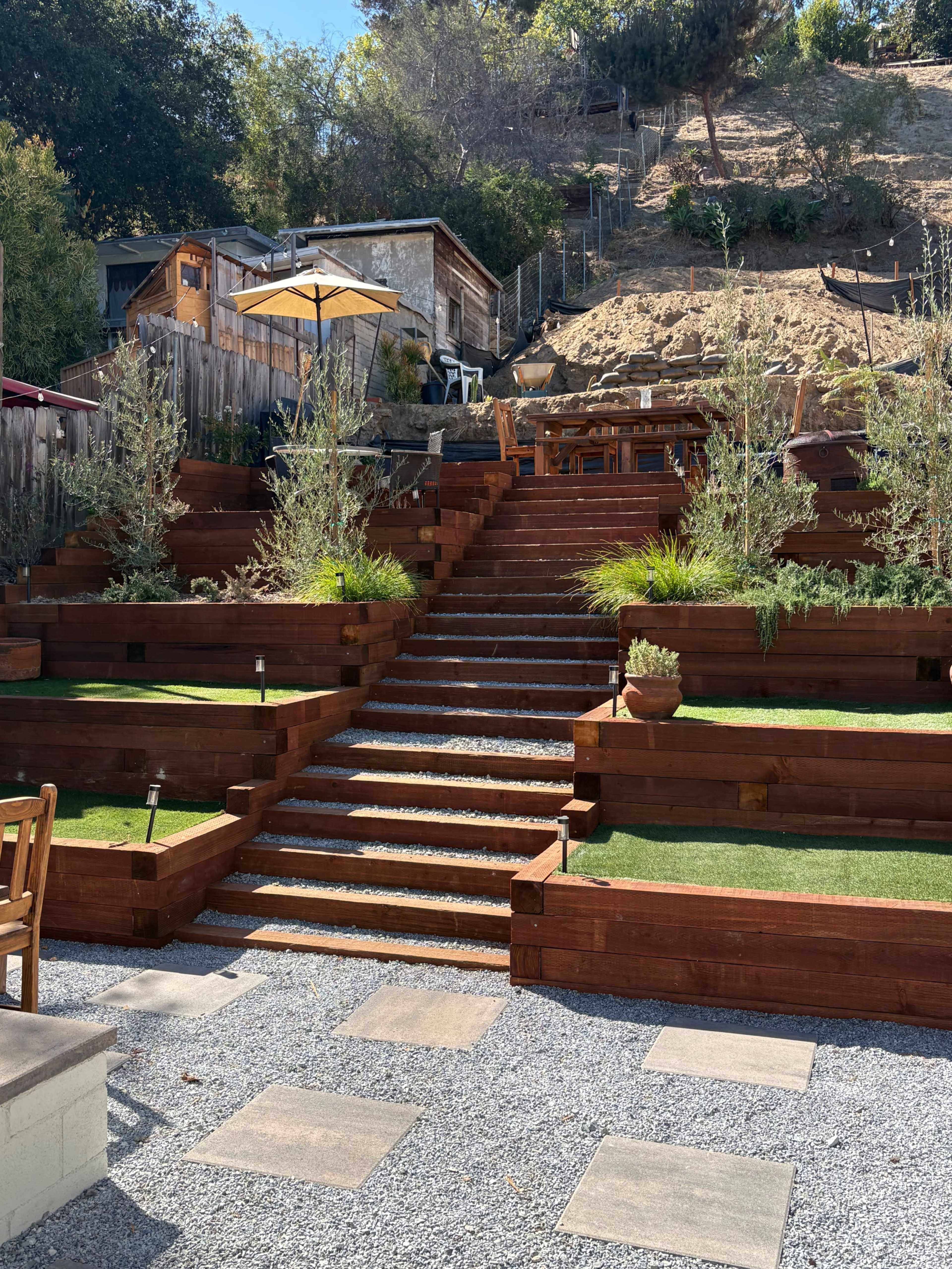 The image shows a terraced garden with wooden steps leading up a hillside, accented by greenery, a dining area, and an umbrella.