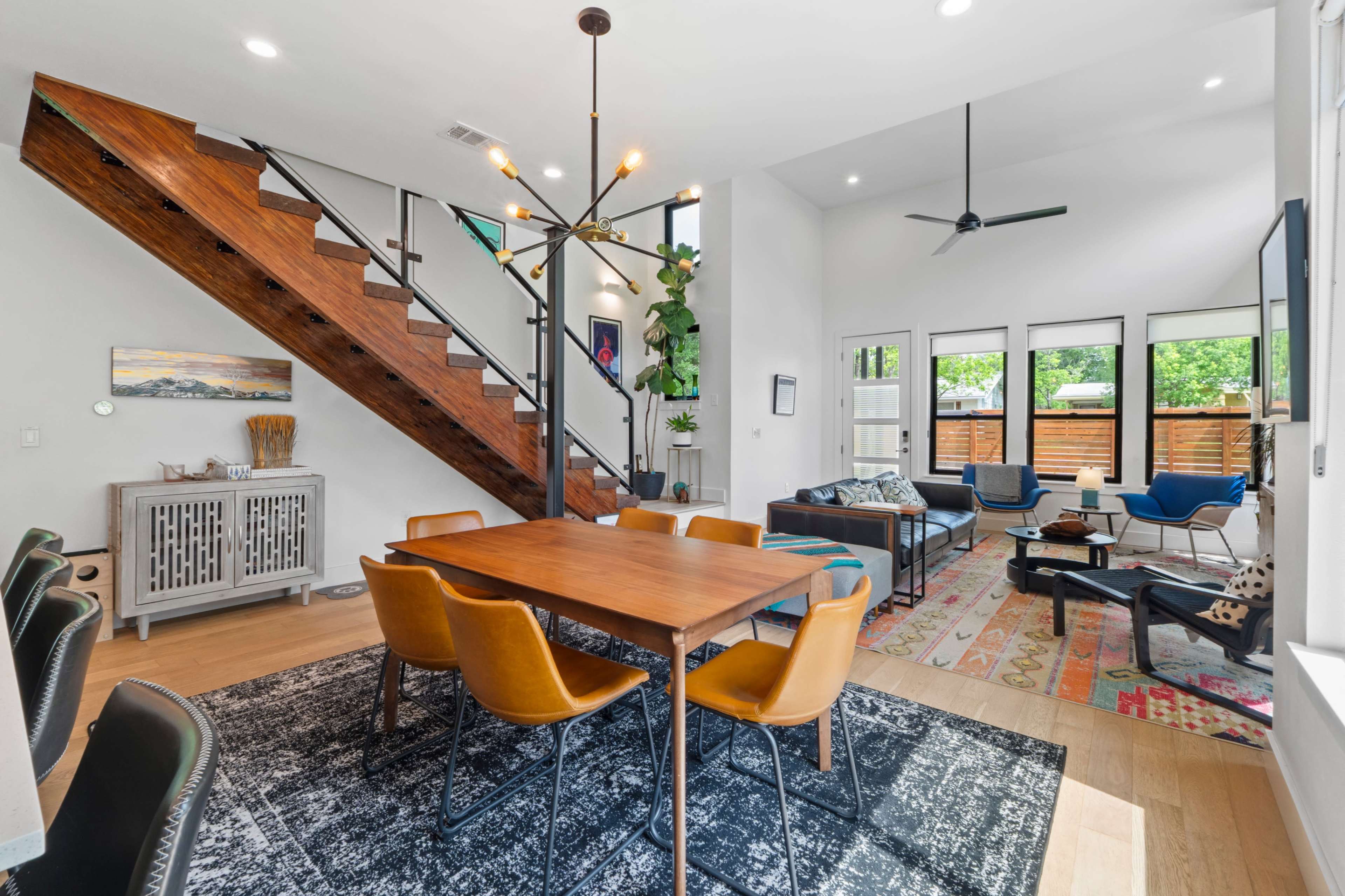 The image shows a modern living space featuring a dining area with a wooden table and orange chairs, a staircase with wooden steps, and a well-furnished sitting area with large windows.